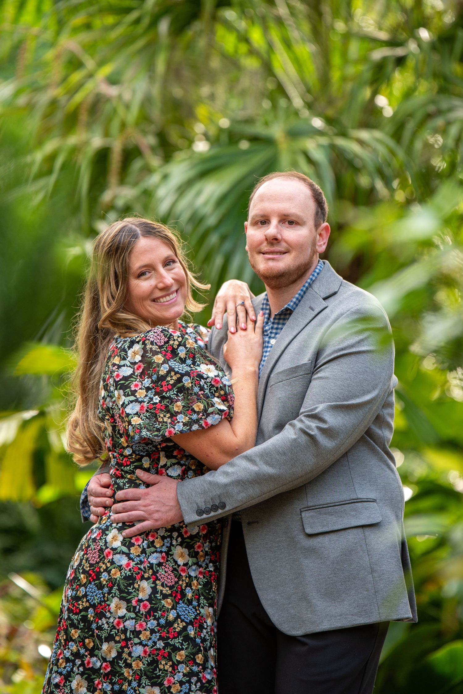 Smiling engaged couple embracing in front of tropical palm foliage during outdoor photo session.