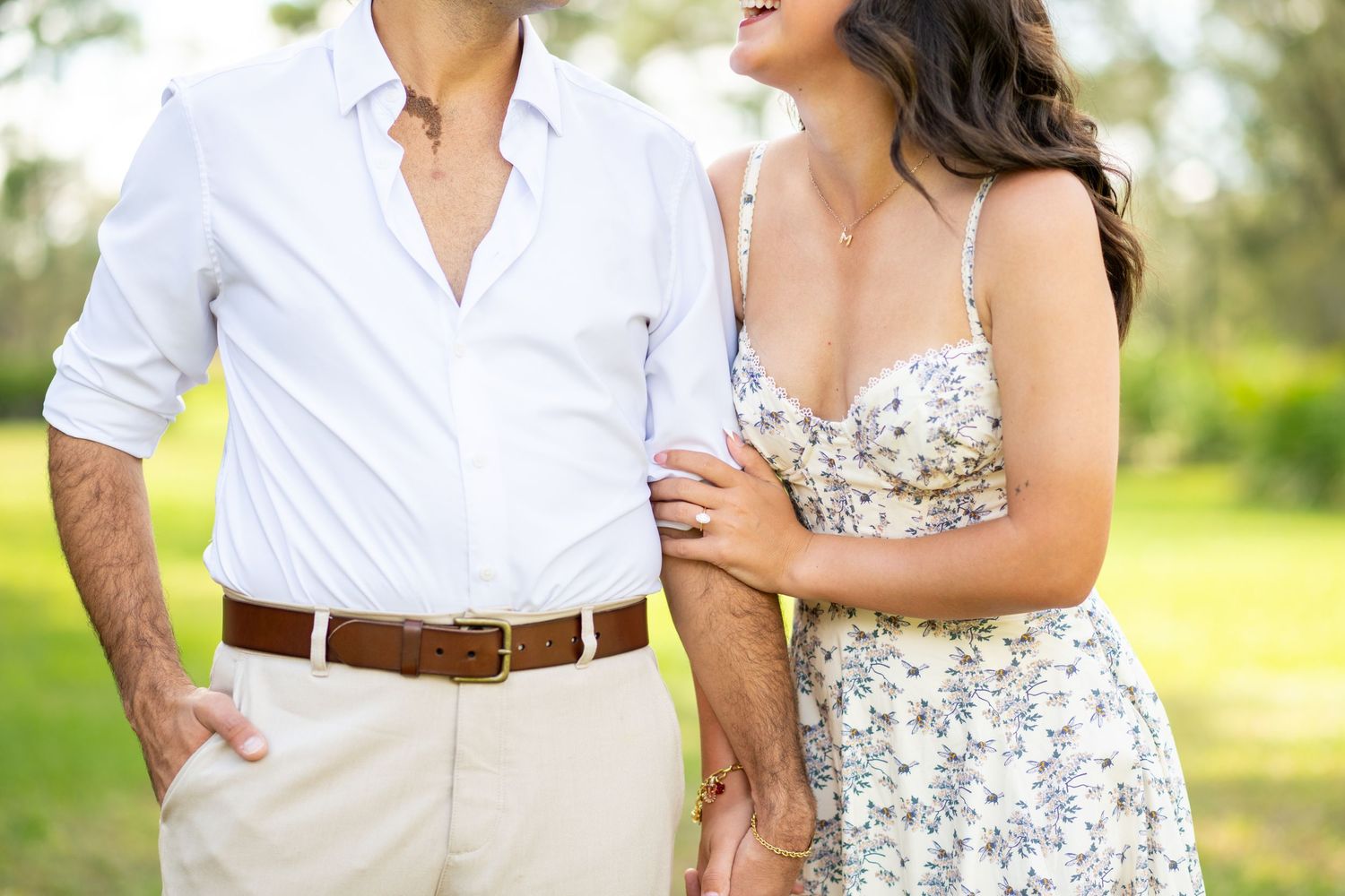 Couple in casual outdoor attire holding hands and laughing together in a sunlit green field.