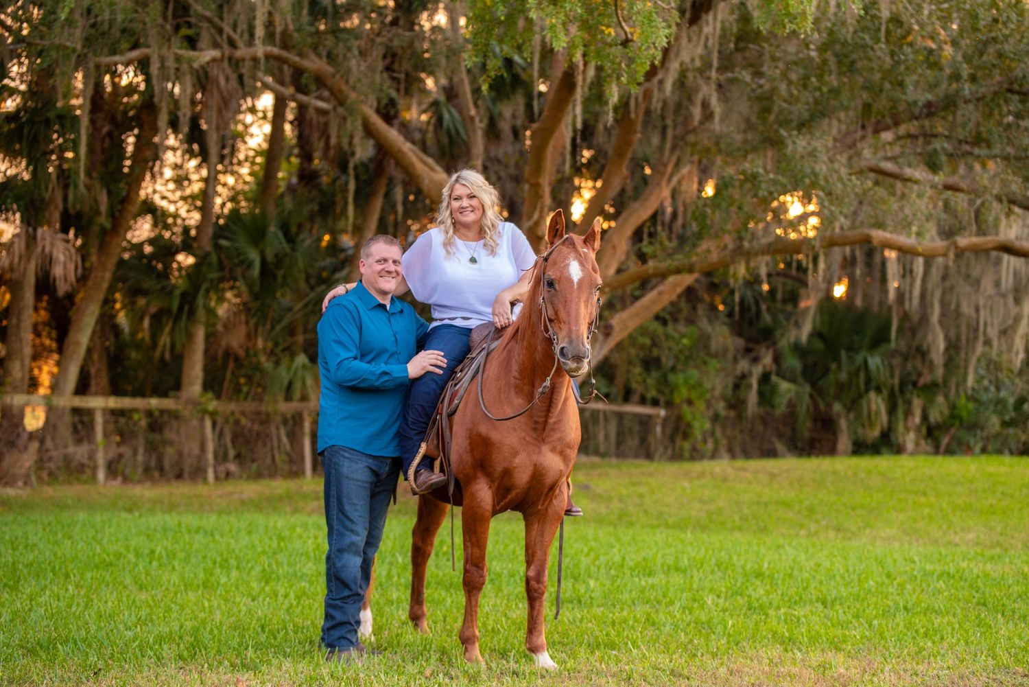 Couple smiling outdoors with a chestnut horse under moss-draped trees at golden hour.