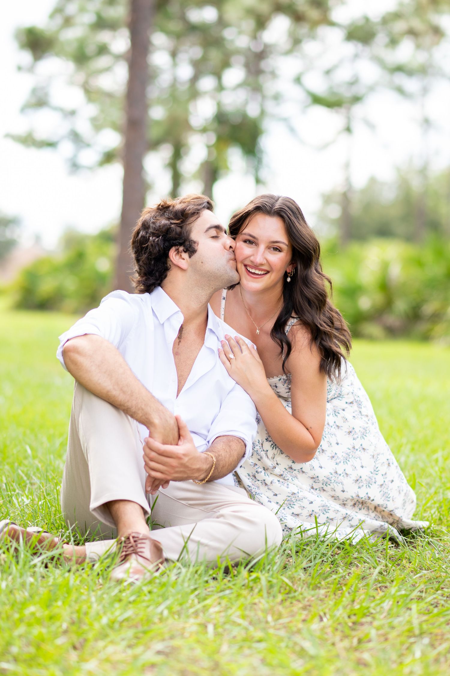 Romantic couple sitting on green grass in a park, man kissing woman on cheek while she smiles happily.