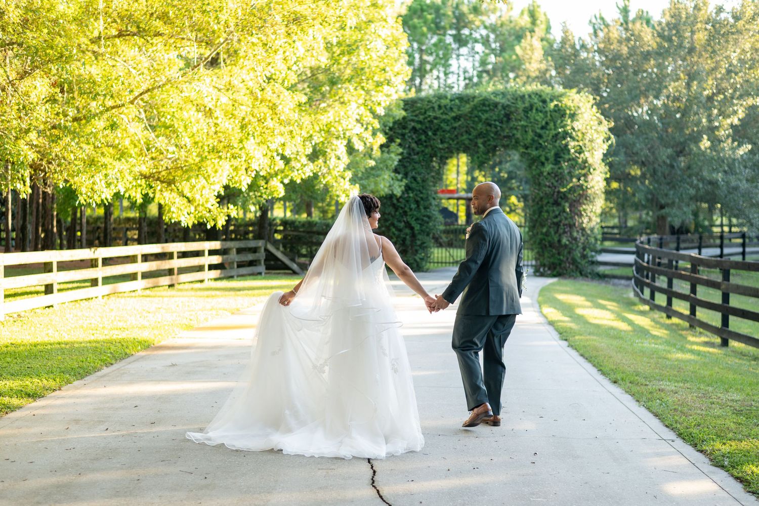 Bride and groom walk hand-in-hand down sunlit path at outdoor farm wedding.