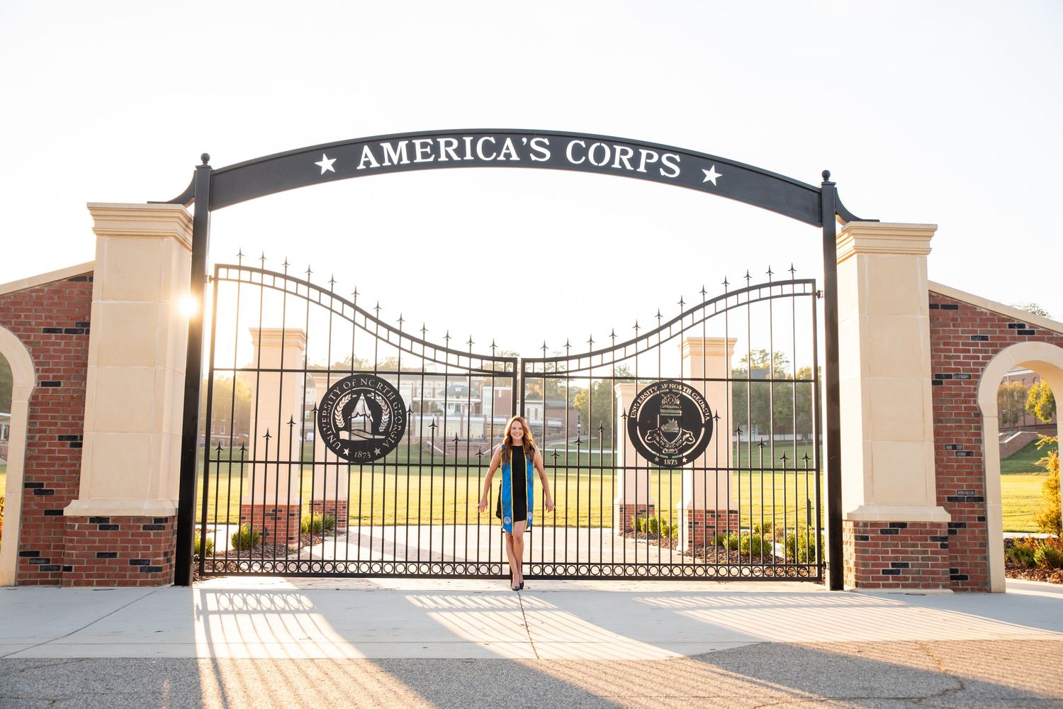 Woman standing before America's Corps ornate iron gate with brick pillars in golden sunset light.