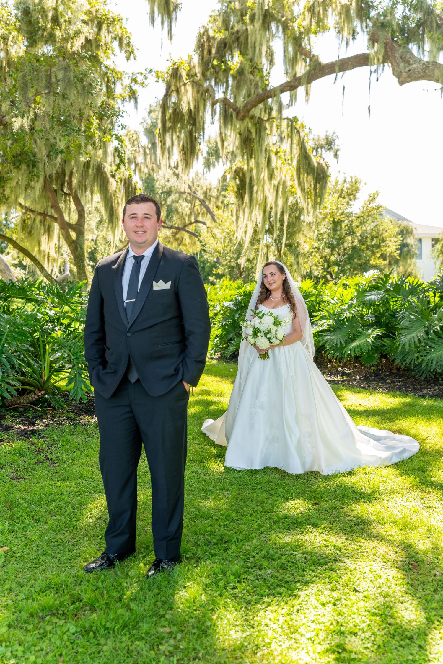 Groom in black suit stands on green lawn as bride in white gown with veil poses behind under Spanish moss trees.