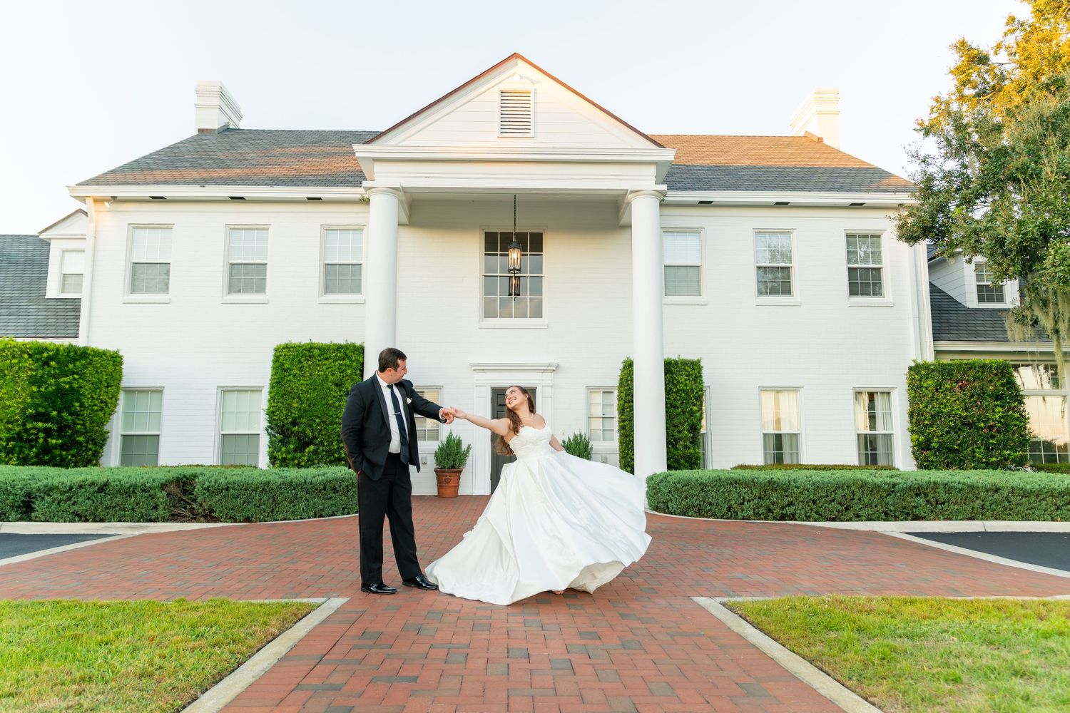 Bride and groom dancing on brick path before white colonial mansion with manicured hedges at outdoor wedding.