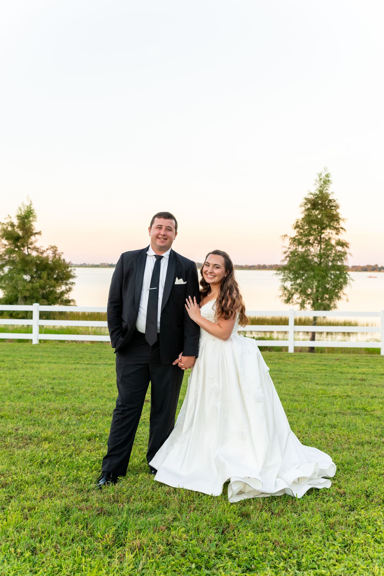 Bride and groom posing together on green lawn at sunset near white fence and lake, bride wearing elegant white ballgown.
