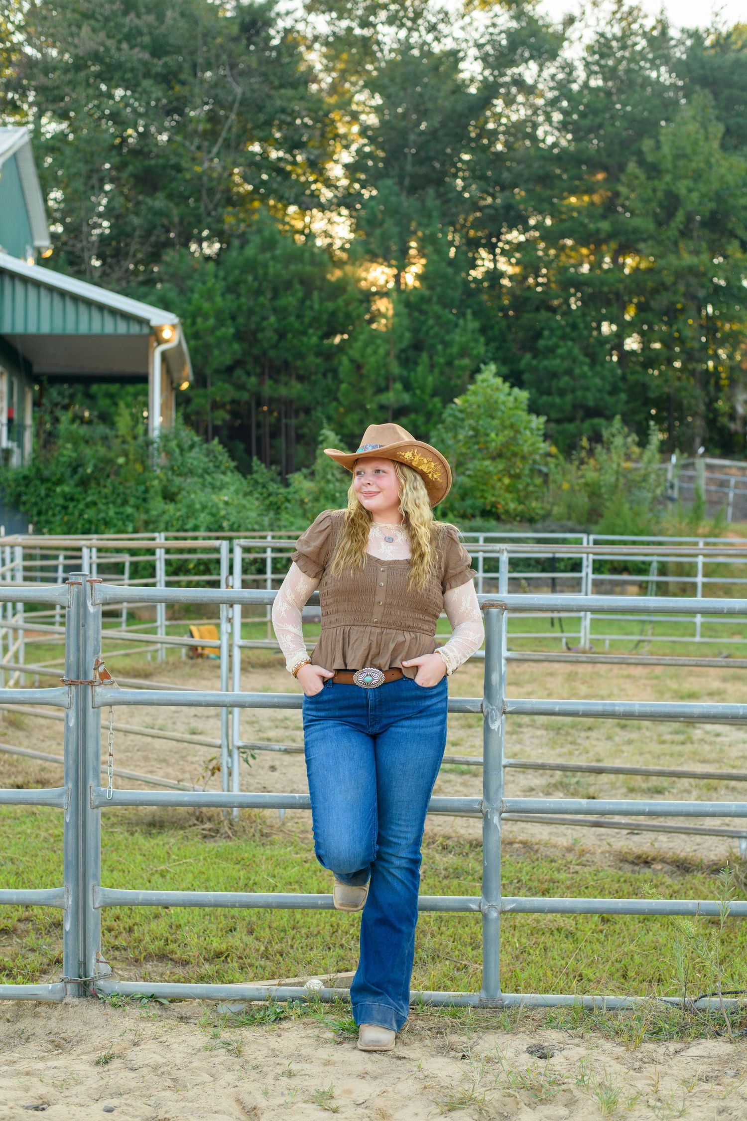 A young woman in a cowboy hat and jeans leans against a metal farm fence, smiling outdoors near a barn.