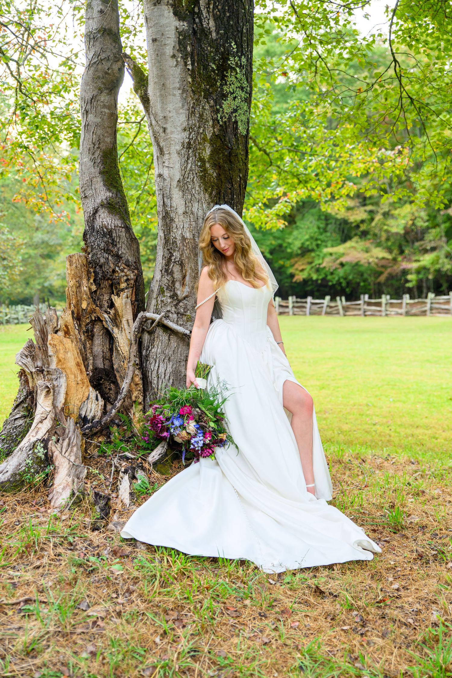 Bride in white gown with slit holds colorful bouquet leaning against large tree in lush green outdoor setting.