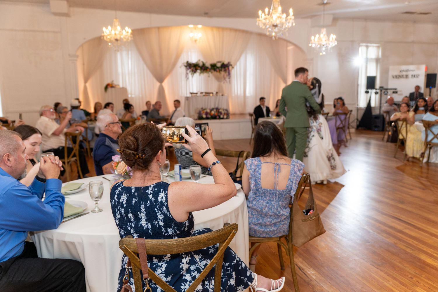 Guests seated at round tables photograph a couple during a wedding reception in an elegant white-draped venue.