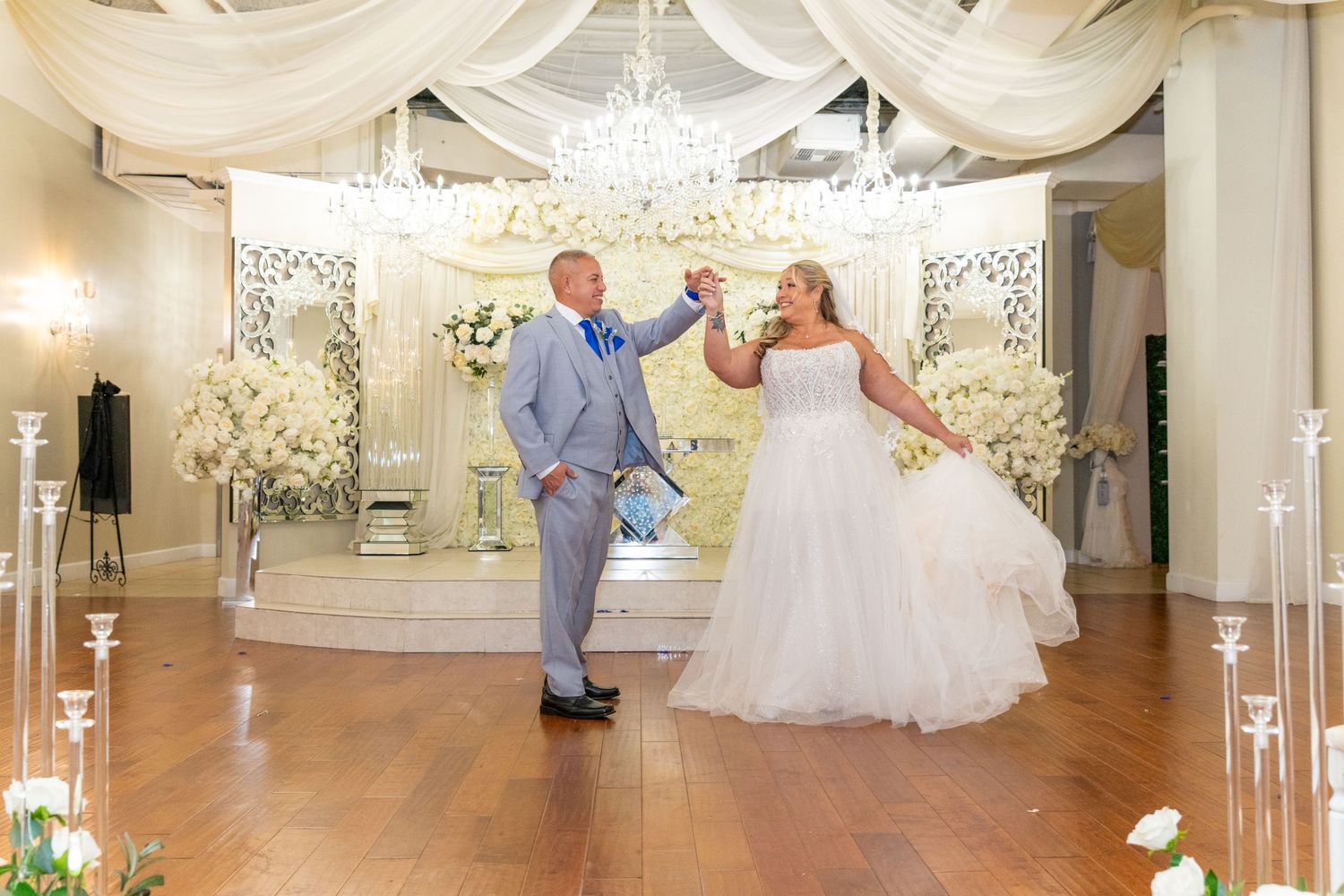 Bride and groom share first dance in elegant white-draped ballroom with crystal chandeliers and floral arrangements.
