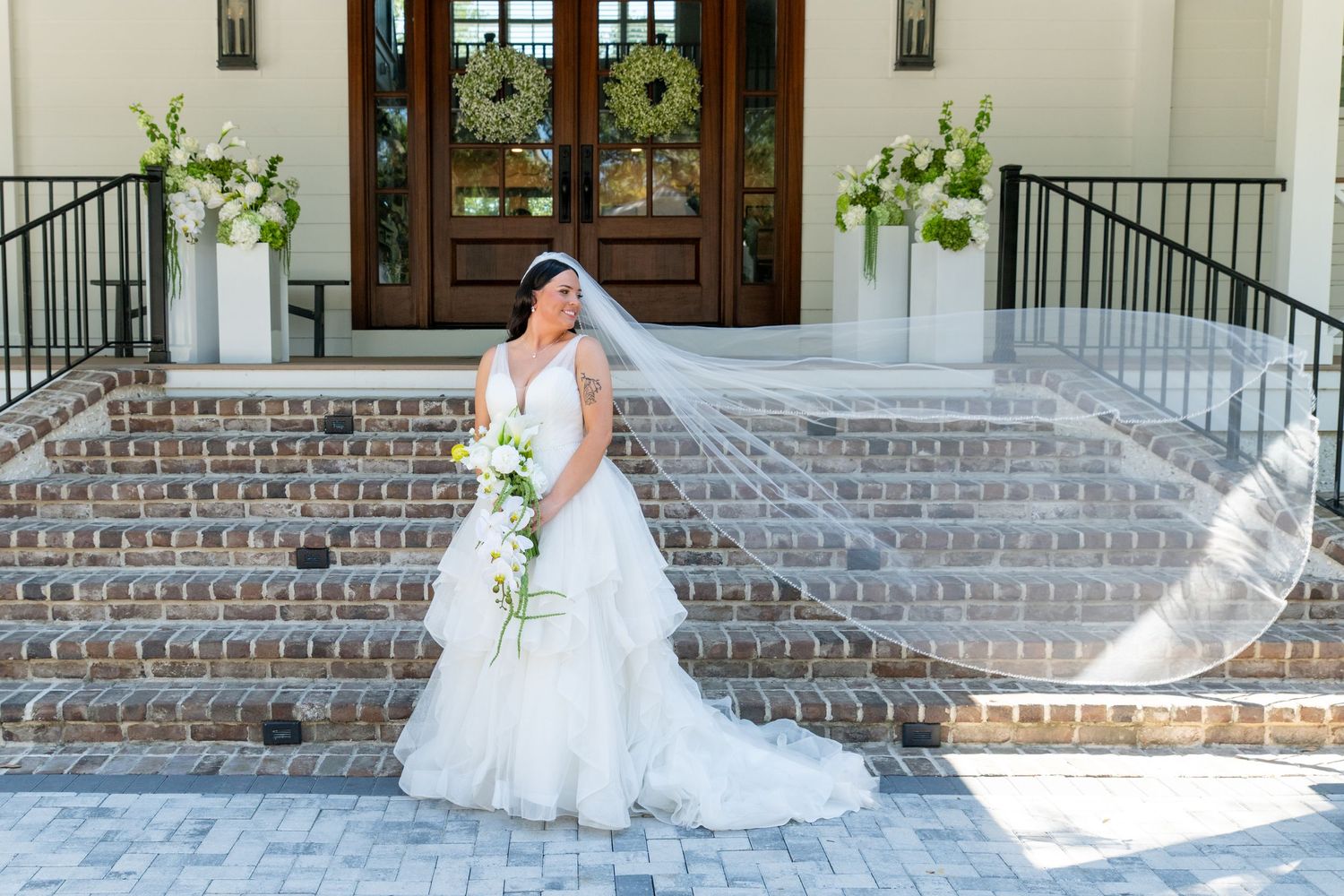 Bride in white gown with flowing veil poses on brick steps before elegant venue doors with floral arrangements.