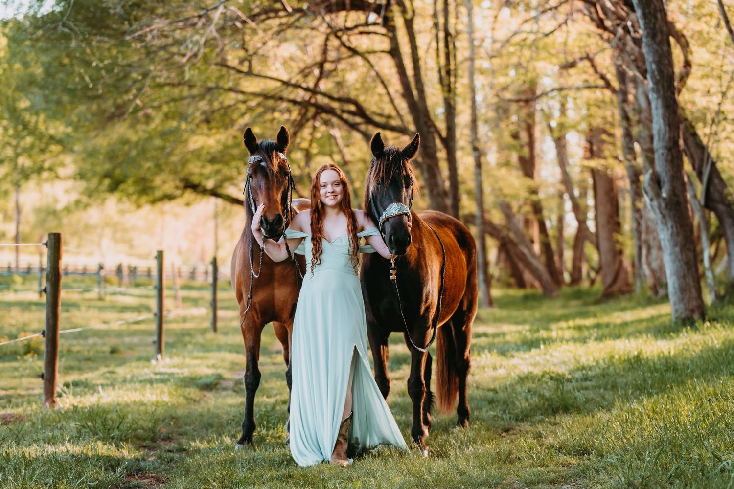 Woman in mint gown poses with two bay horses in a sunlit green pasture surrounded by trees.