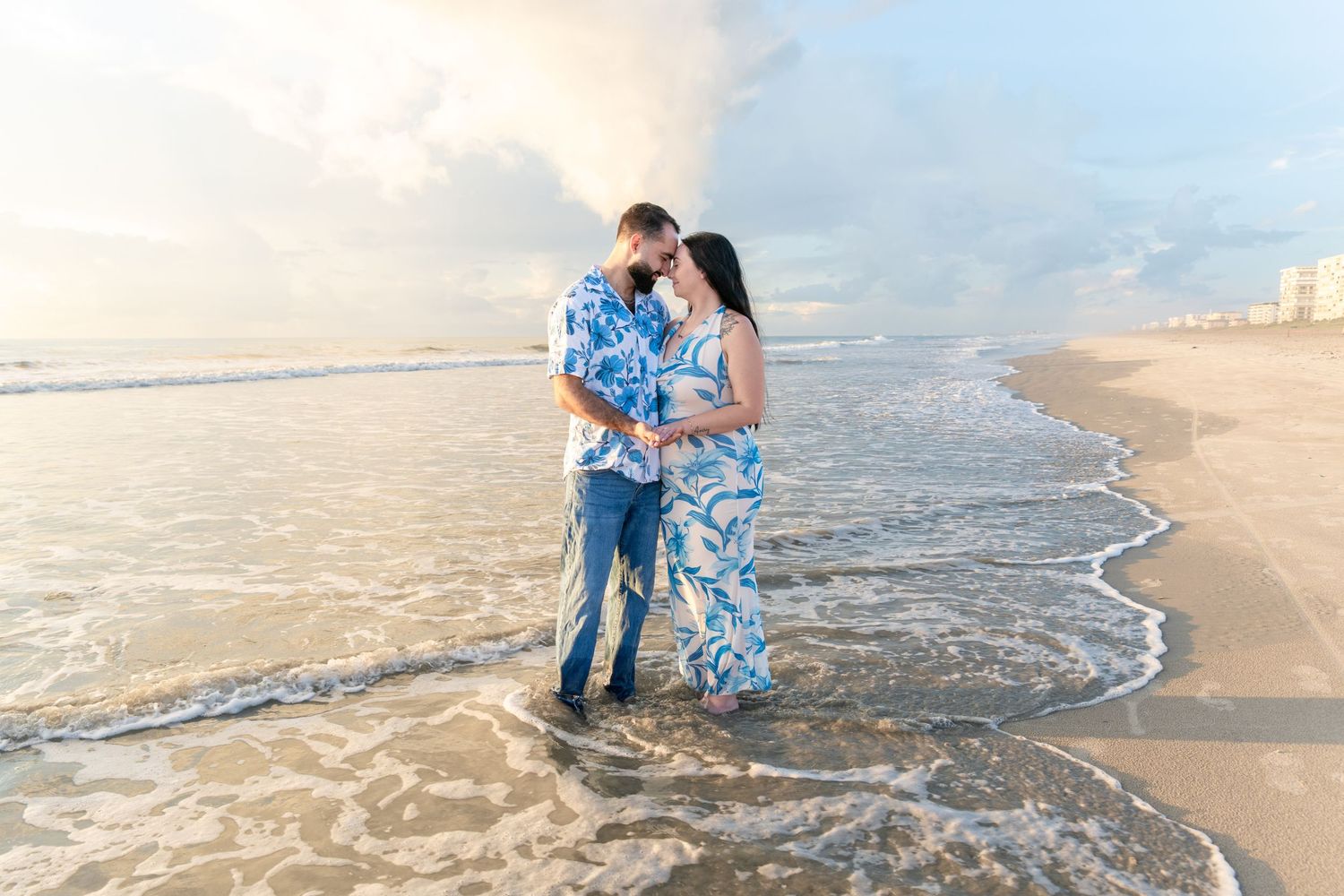 Couple in matching blue outfits standing in ocean waves on a sandy beach at golden hour.