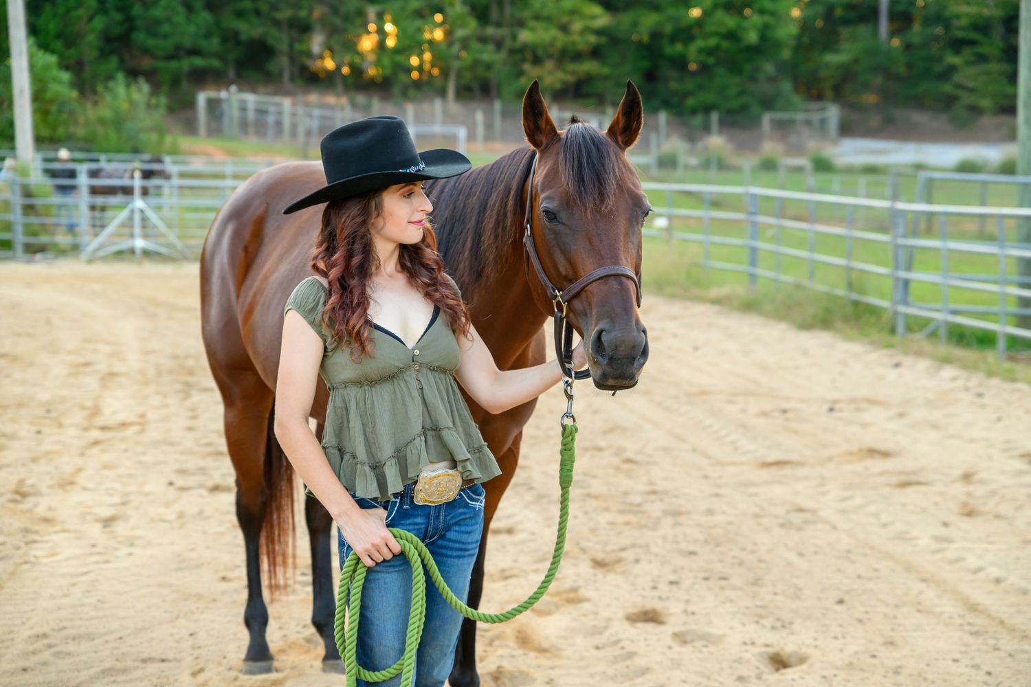 Young woman in cowboy hat and green top leads a chestnut horse by green rope in sandy ranch corral.