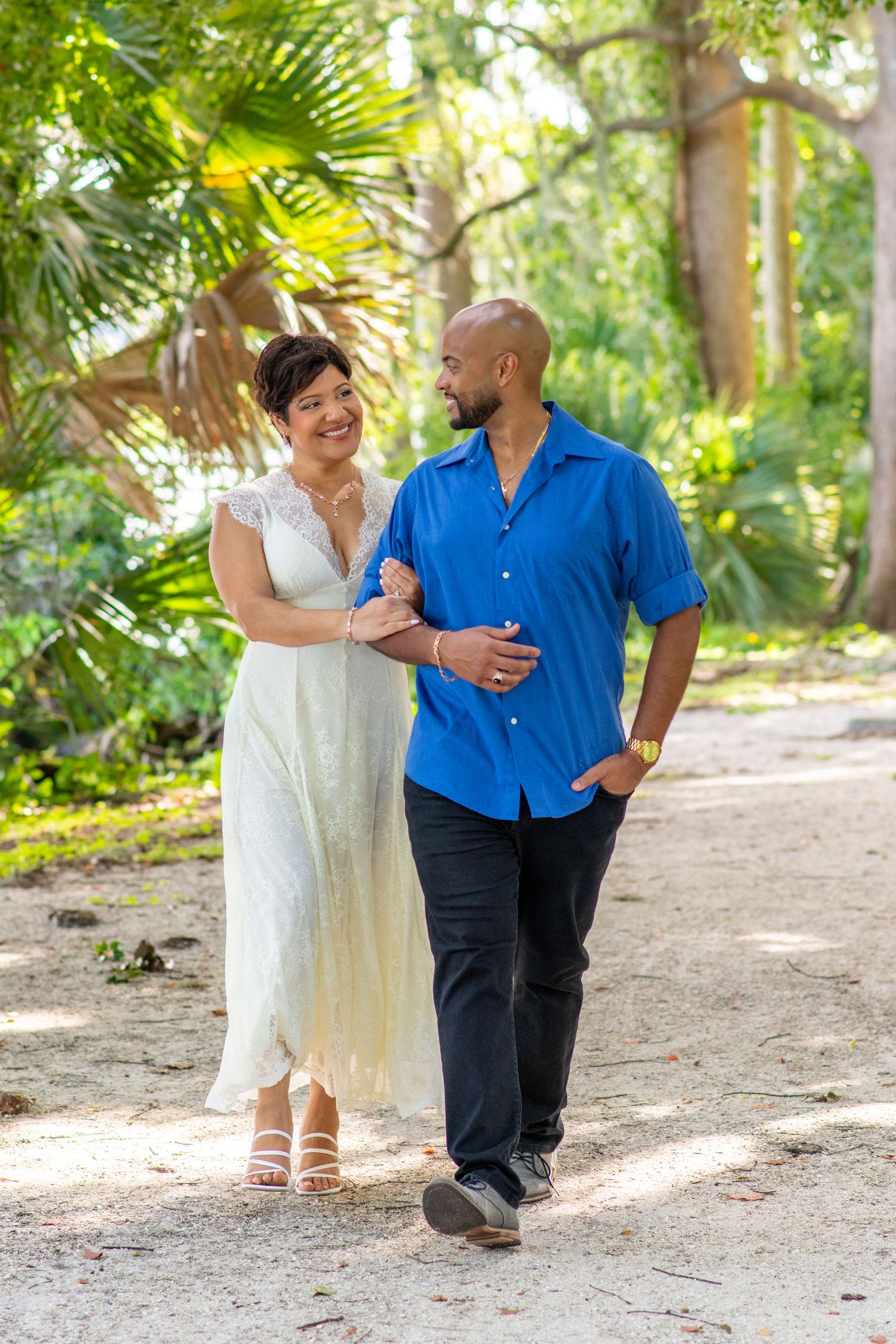 Couple walking hand in hand on a sunny path surrounded by lush tropical greenery, woman in white dress, man in blue shirt.