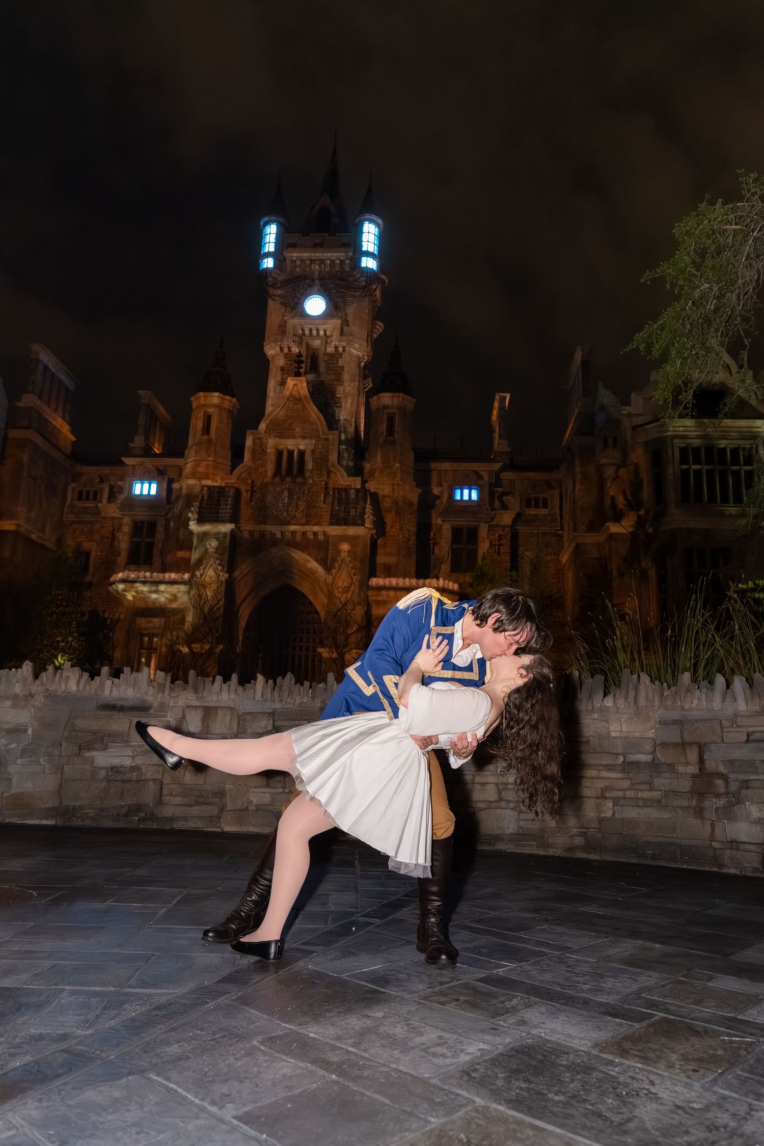 Couple sharing a romantic dip kiss at night in front of an illuminated gothic castle.