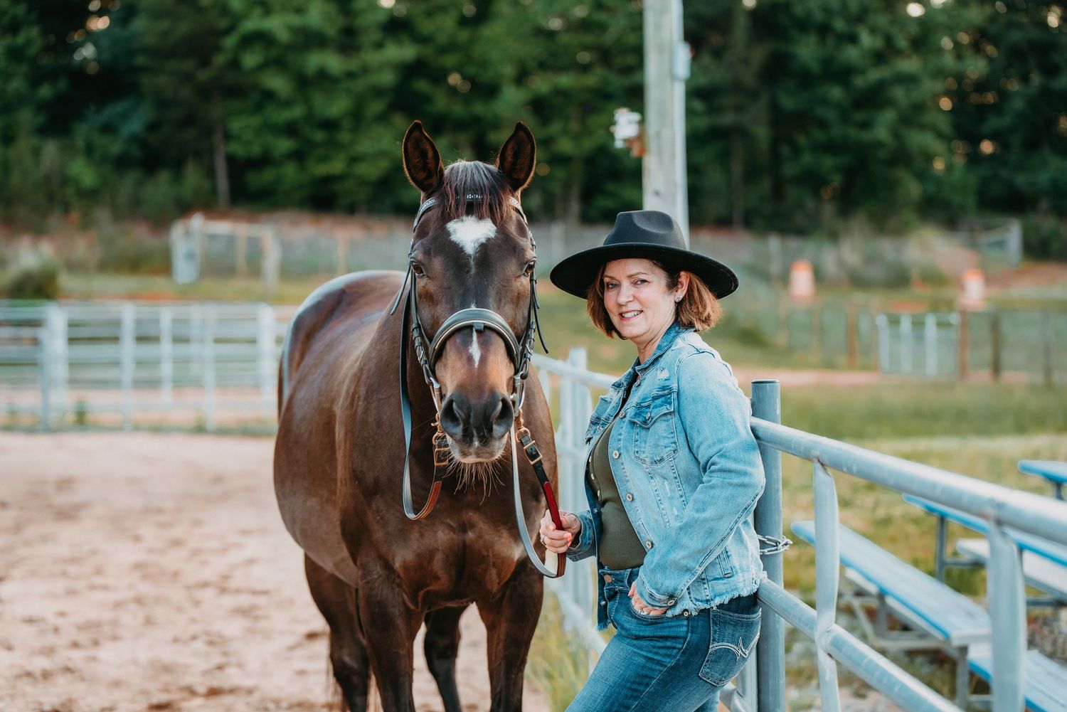 Woman in cowboy hat and denim jacket smiling beside a brown horse in a fenced equestrian arena.