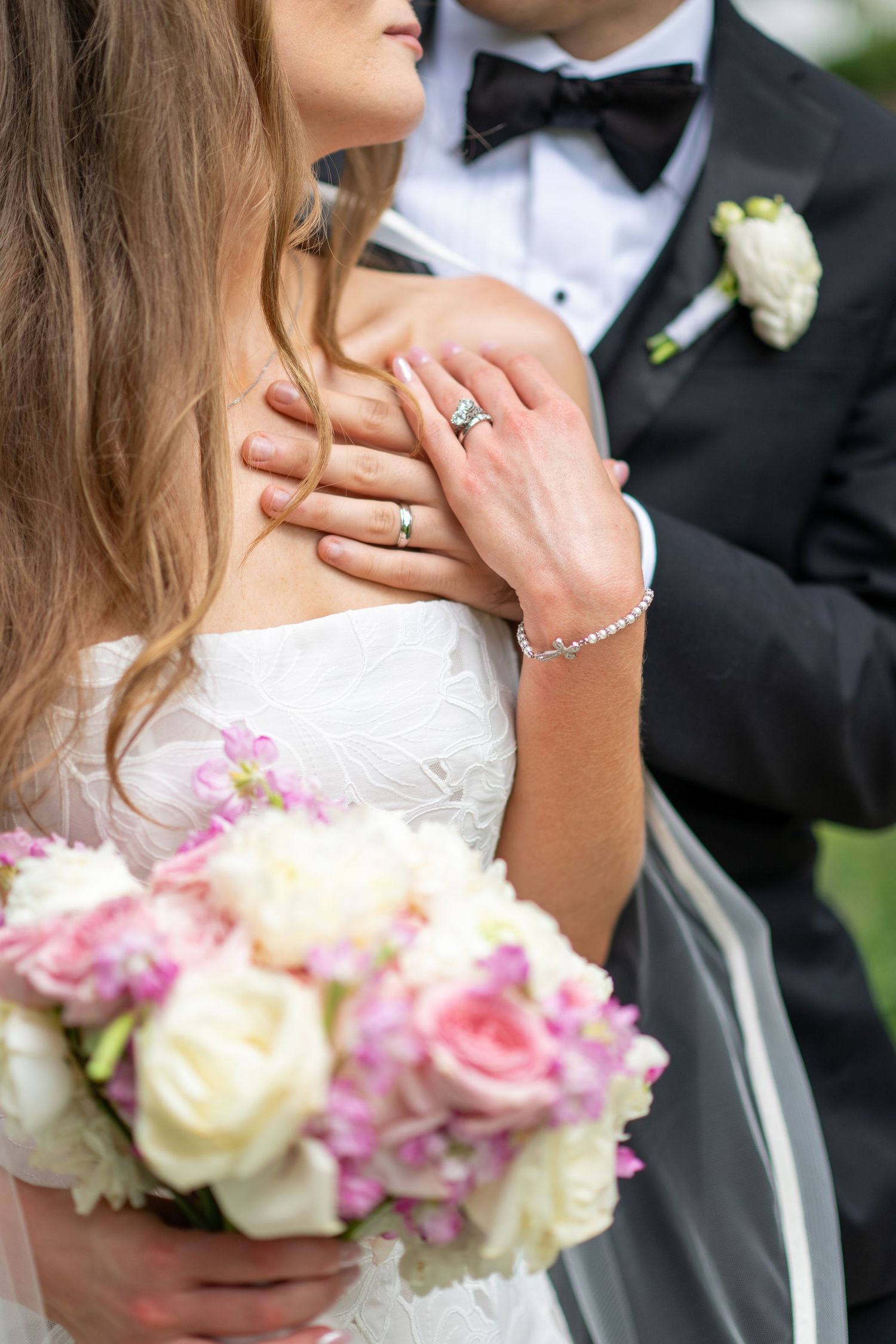 Bride holding pink and white bouquet with groom's hand on her shoulder, both wearing wedding rings.