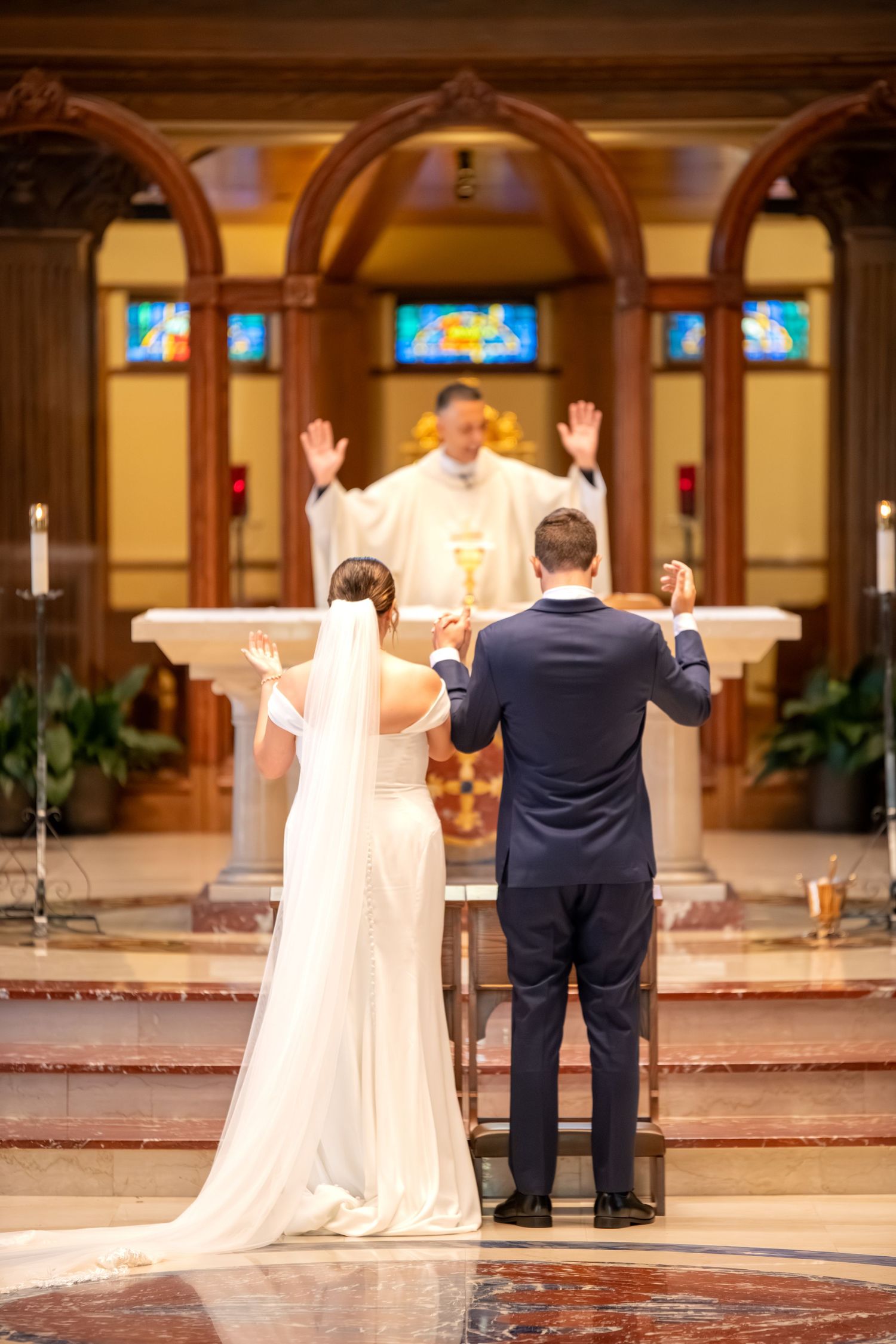 Bride and groom raise hands at church altar during wedding ceremony with priest officiating before stained glass windows.