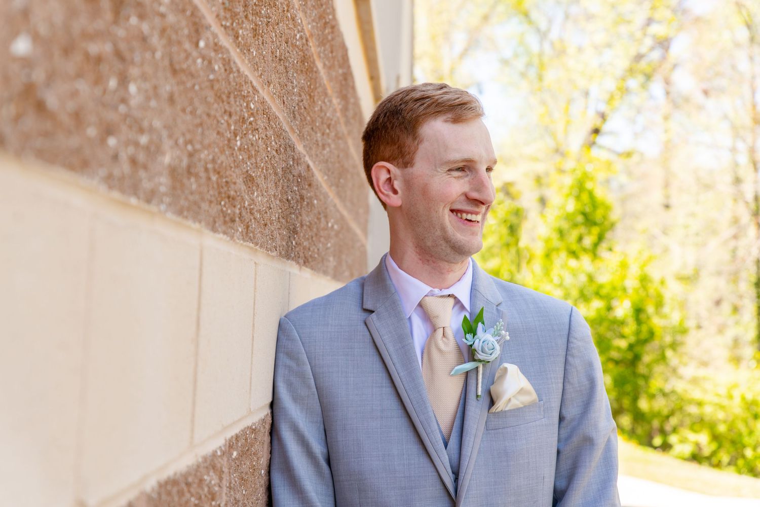 Smiling groom in gray suit with boutonniere leans against stone wall outdoors on sunny wedding day.