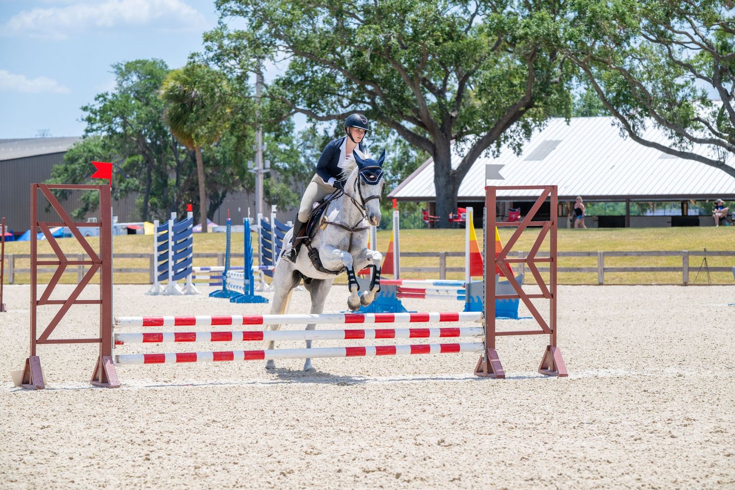 Gray horse and rider jumping over red and white fence in outdoor show jumping competition.