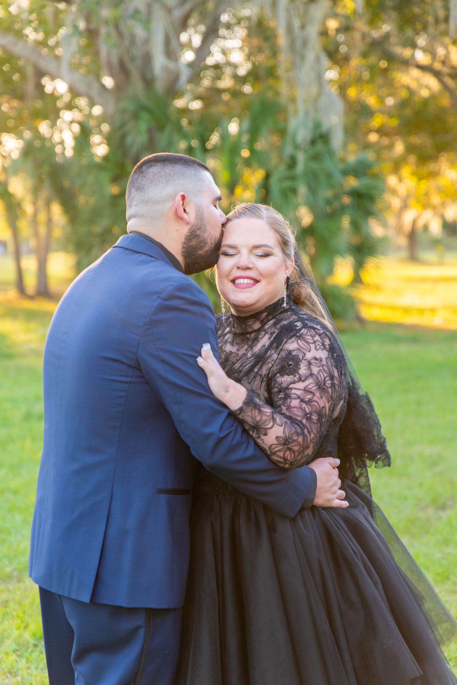 Groom in navy suit kisses smiling bride wearing black lace wedding dress outdoors.