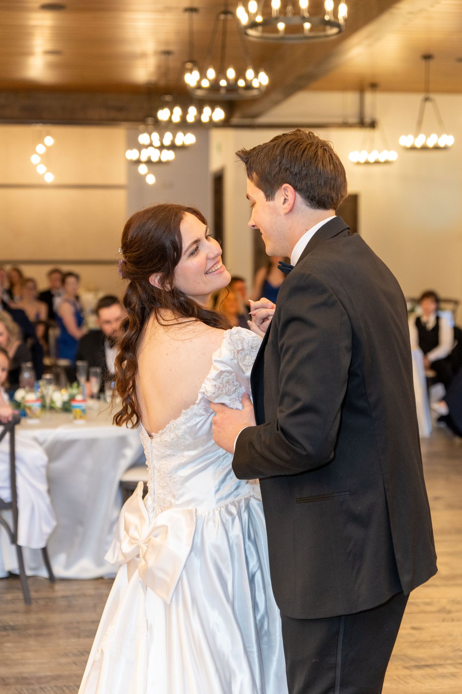 Bride and groom share their first dance at wedding reception, bride in white gown smiling at groom in black tuxedo.