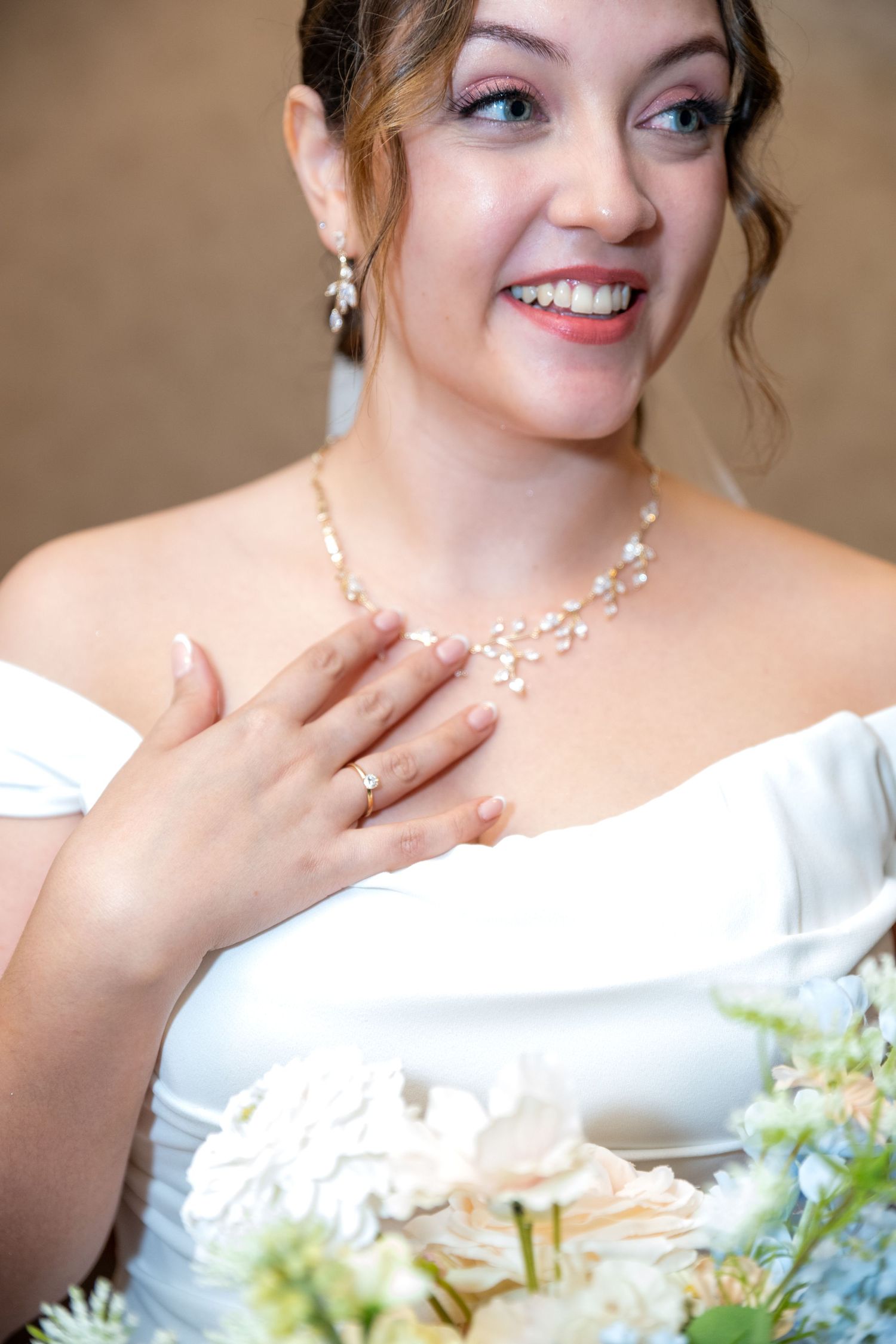 Smiling bride in white off-shoulder gown holds bouquet, wearing pearl necklace and earrings with wedding ring.