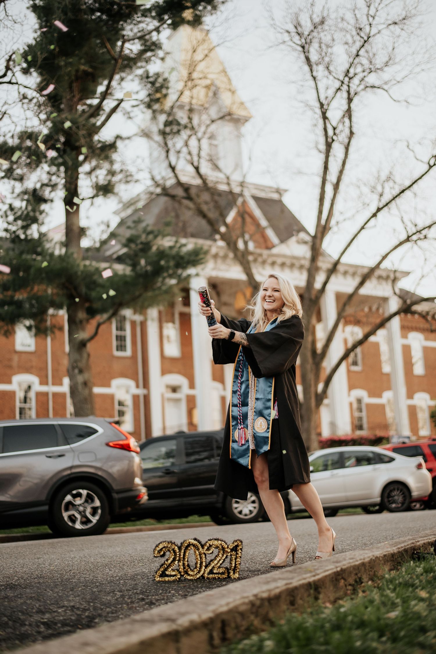 Graduate in cap and gown celebrates popping champagne near 2021 balloon numbers on campus.