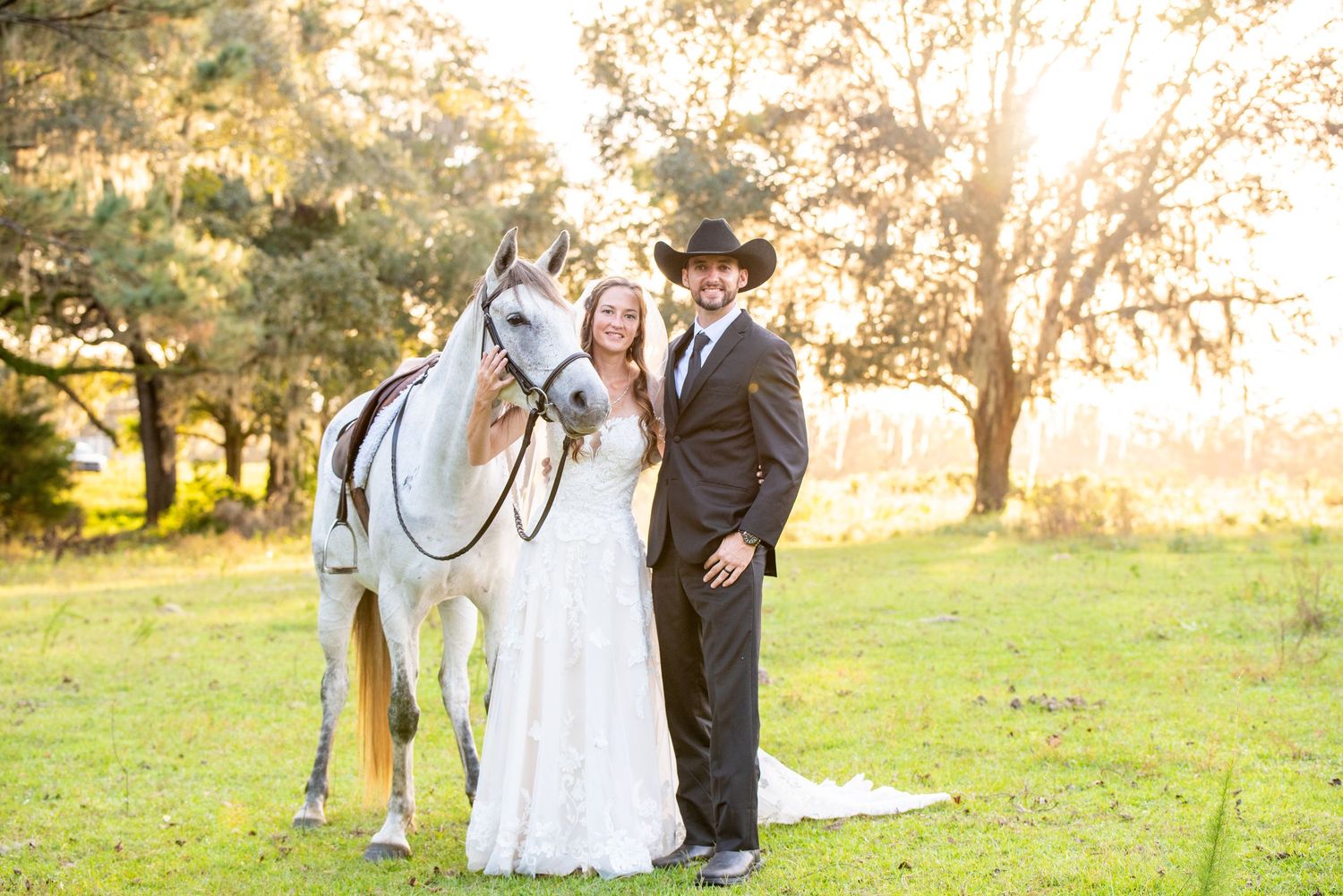 Bride and groom in cowboy hat posing with white horse on green field at golden hour outdoor wedding.