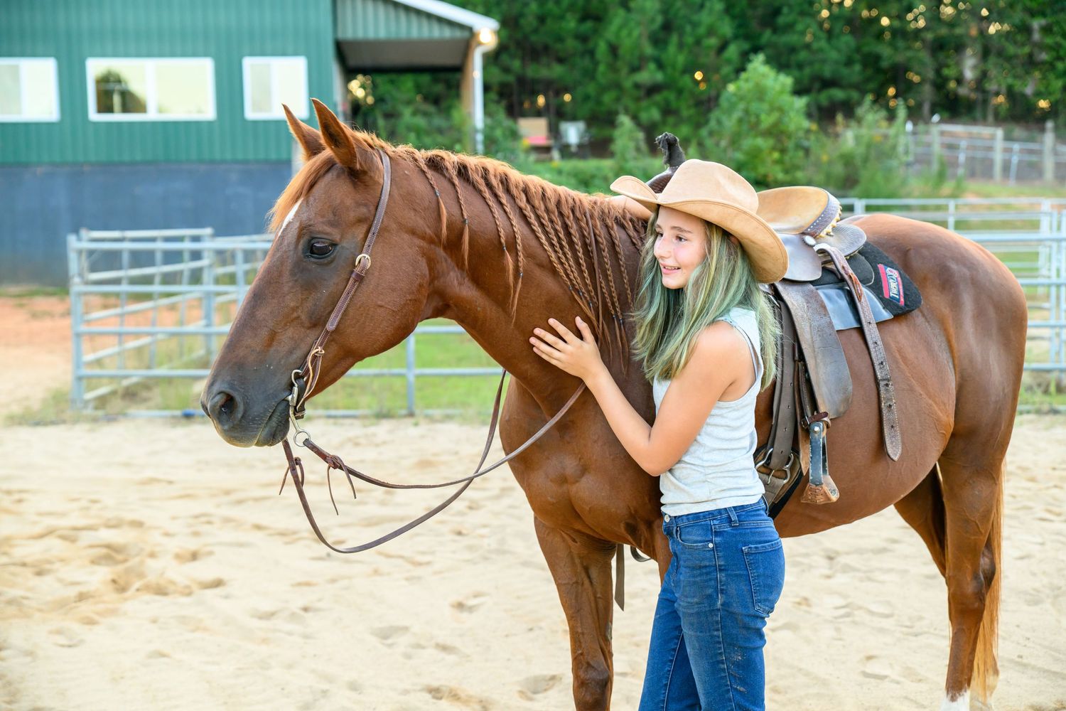 Young blonde woman in cowboy hat gently petting a chestnut horse with western saddle in sandy ranch corral.