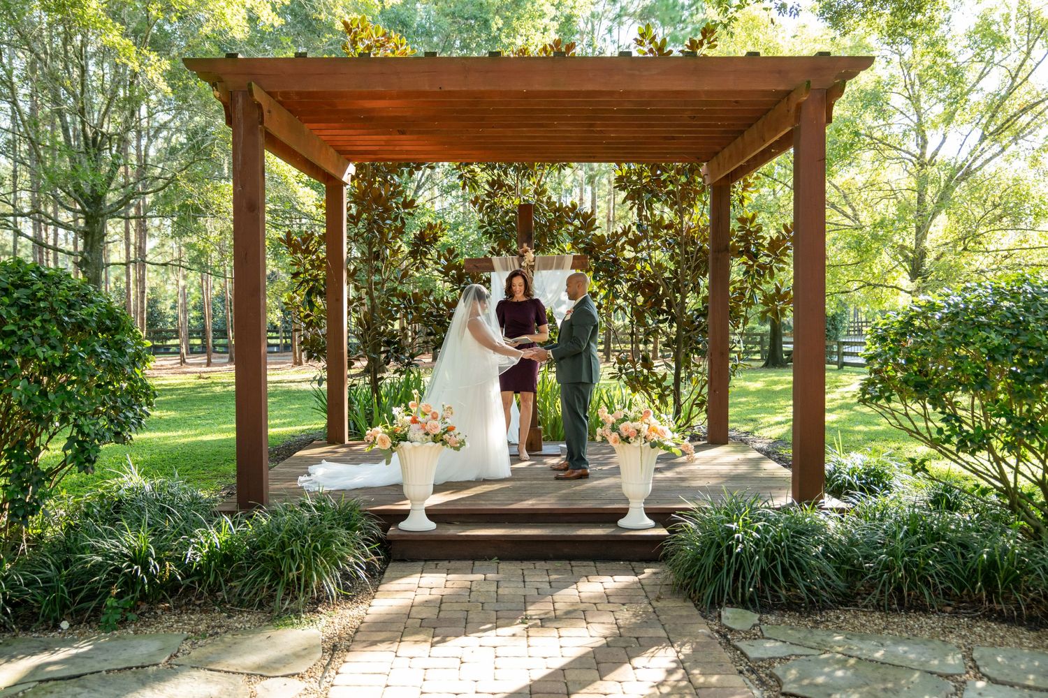Couple exchanges vows under a wooden pergola surrounded by lush greenery and floral arrangements in an outdoor garden wedding.