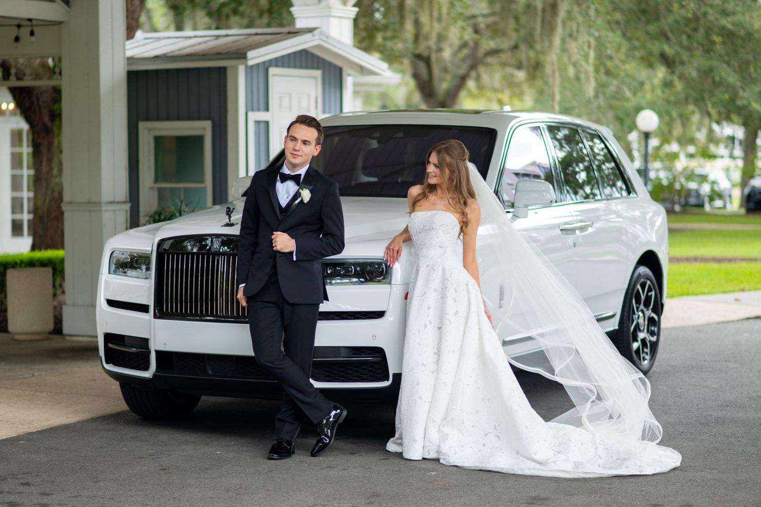 Bride and groom posing elegantly beside a white Rolls-Royce SUV at an outdoor wedding venue.