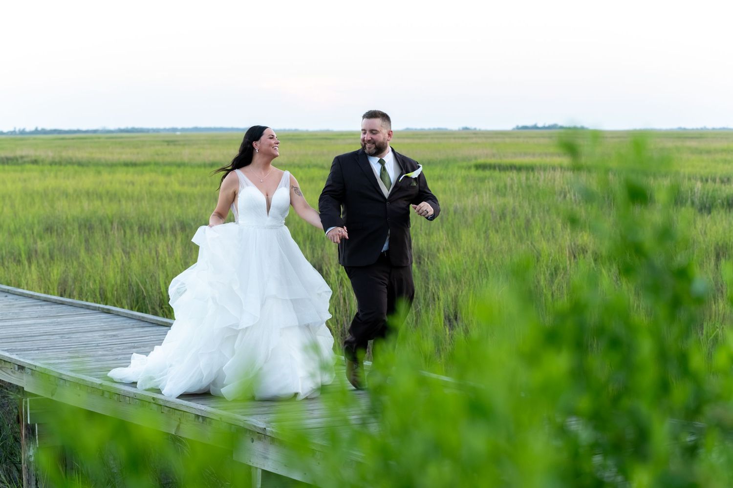 Bride and groom run hand-in-hand on a wooden boardwalk through lush green marshlands on their wedding day.