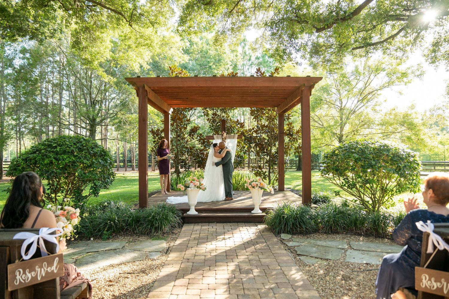 Couple kissing under wooden pergola at outdoor garden wedding ceremony with lush green trees and seated guests.