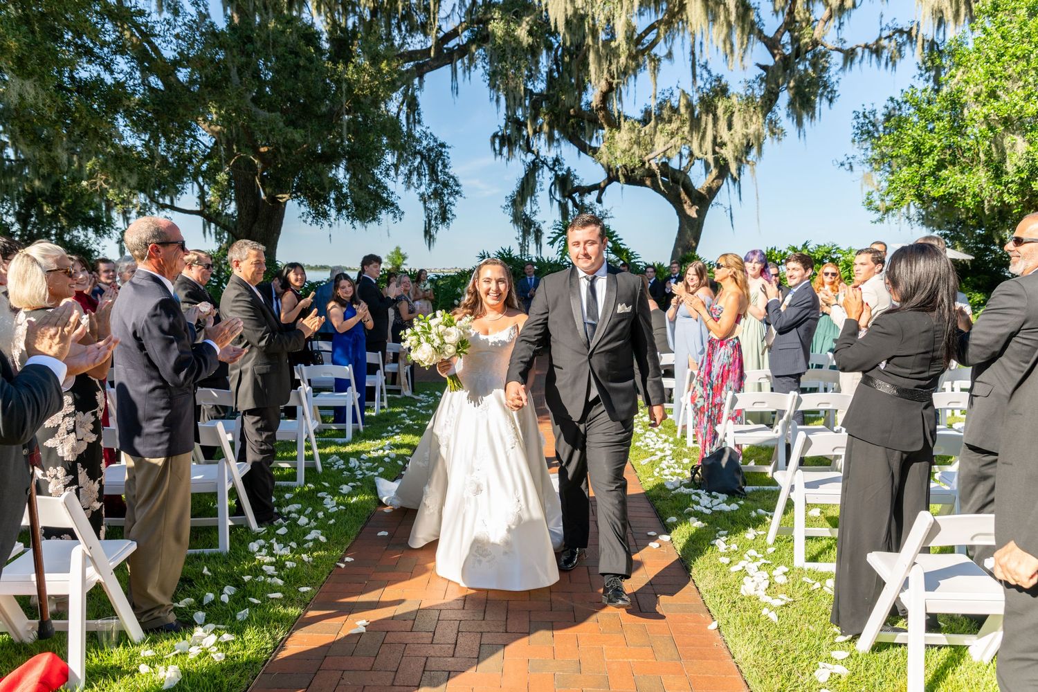 Bride and groom walk down outdoor aisle after wedding ceremony, guests cheering, white chairs, Spanish moss trees.