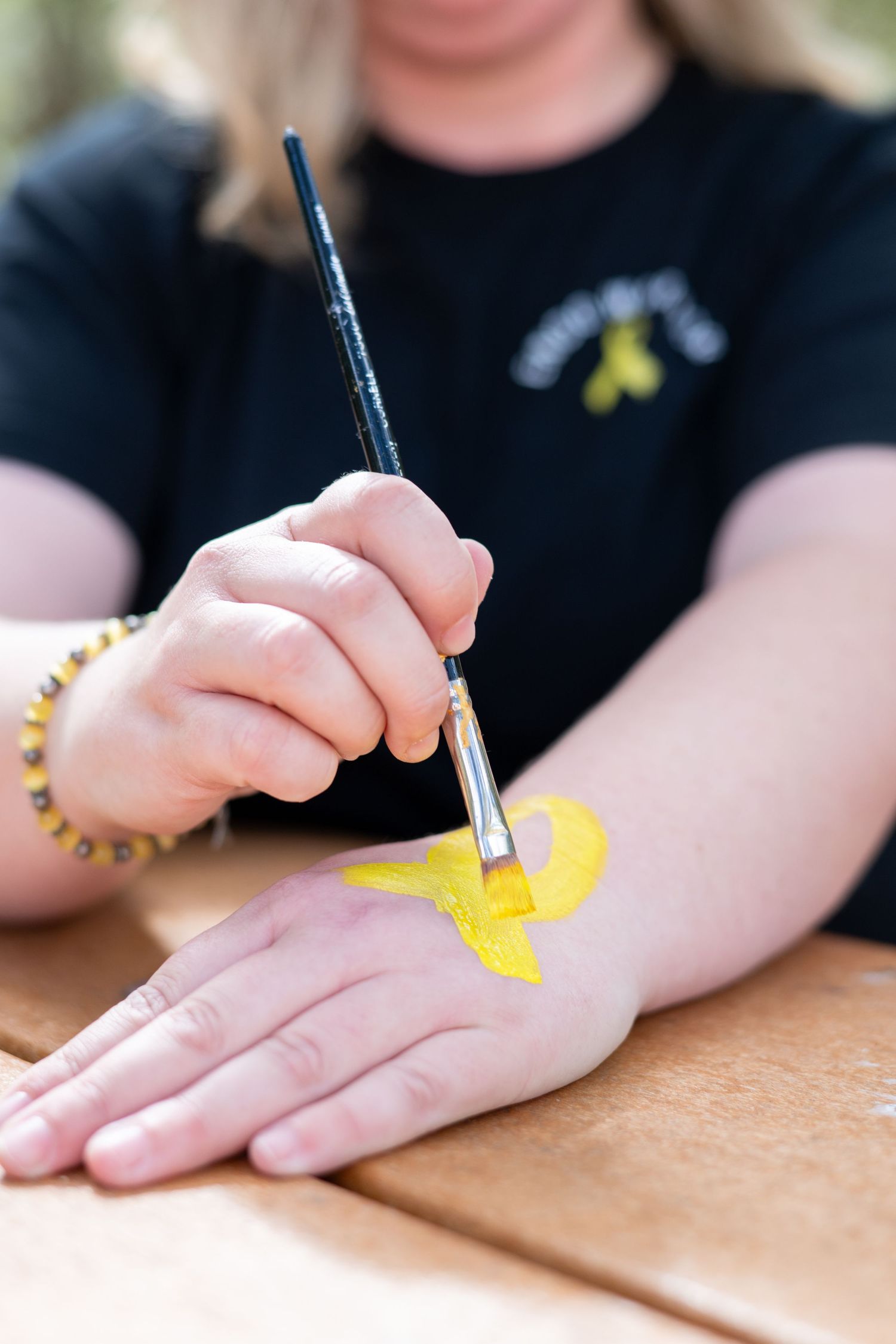 Woman painting a yellow ribbon on her hand with a brush, symbolizing childhood cancer awareness.