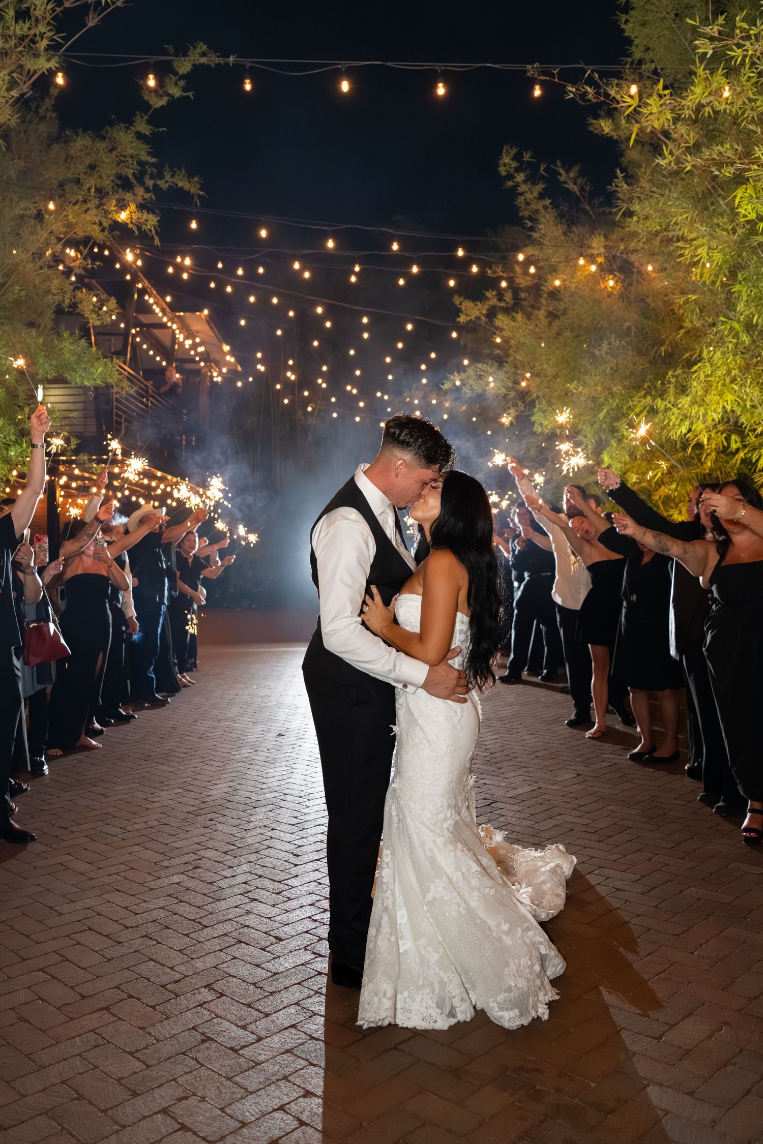 Bride and groom share a kiss during a romantic sparkler send-off under string lights at an outdoor night wedding.