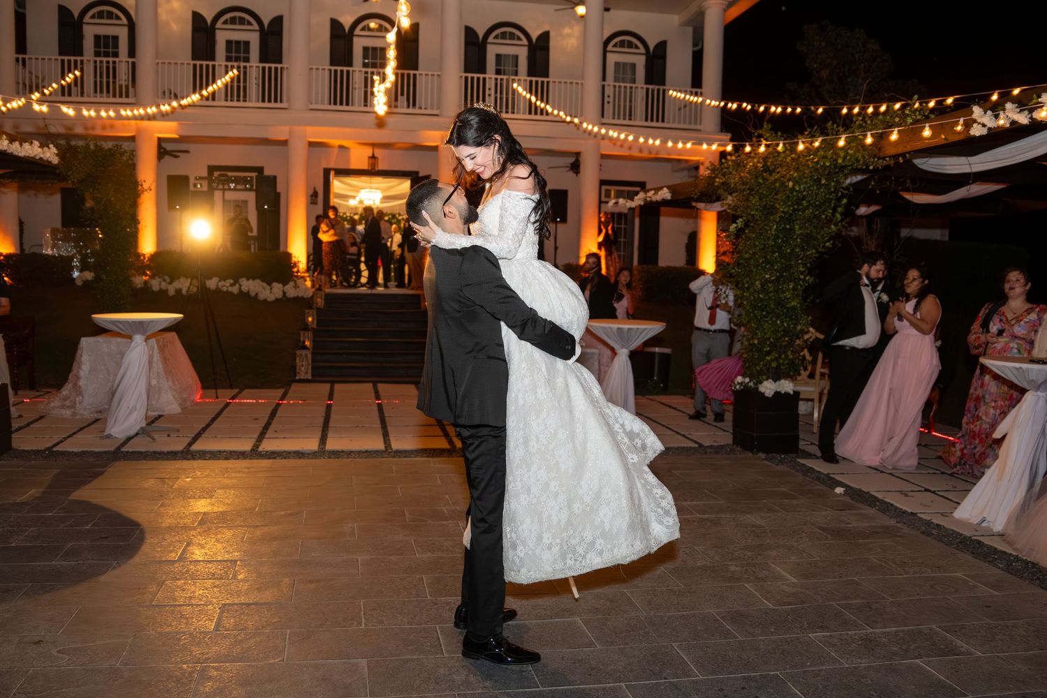 Groom lifts bride during their first dance at an outdoor wedding reception at night.