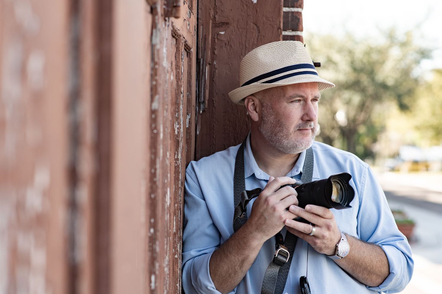 Middle-aged male photographer in straw hat holding camera against rustic red brick wall outdoors.