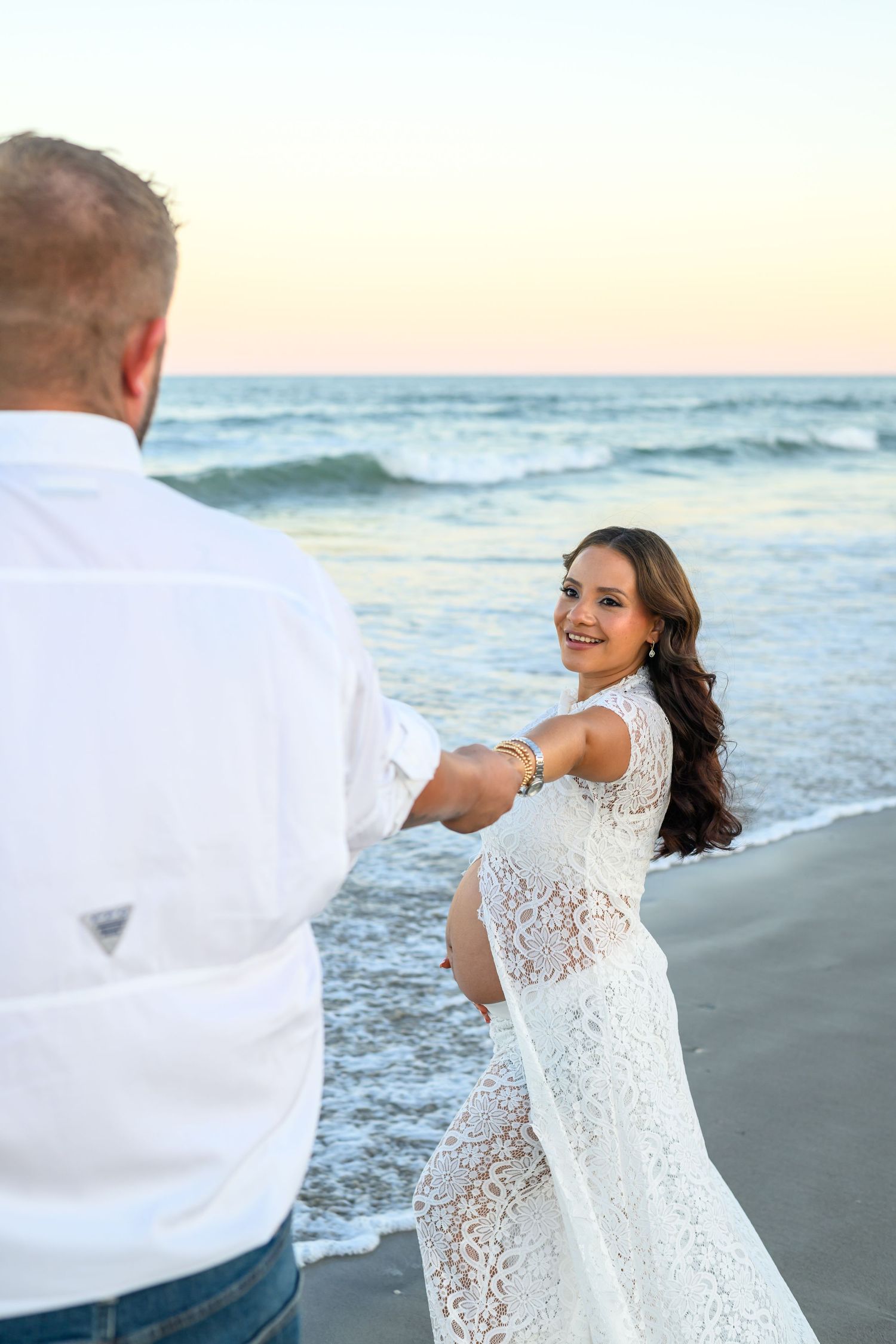 Pregnant woman in a white lace dress smiles while holding a man's hand on a sandy beach at sunset, with ocean waves behind them.
