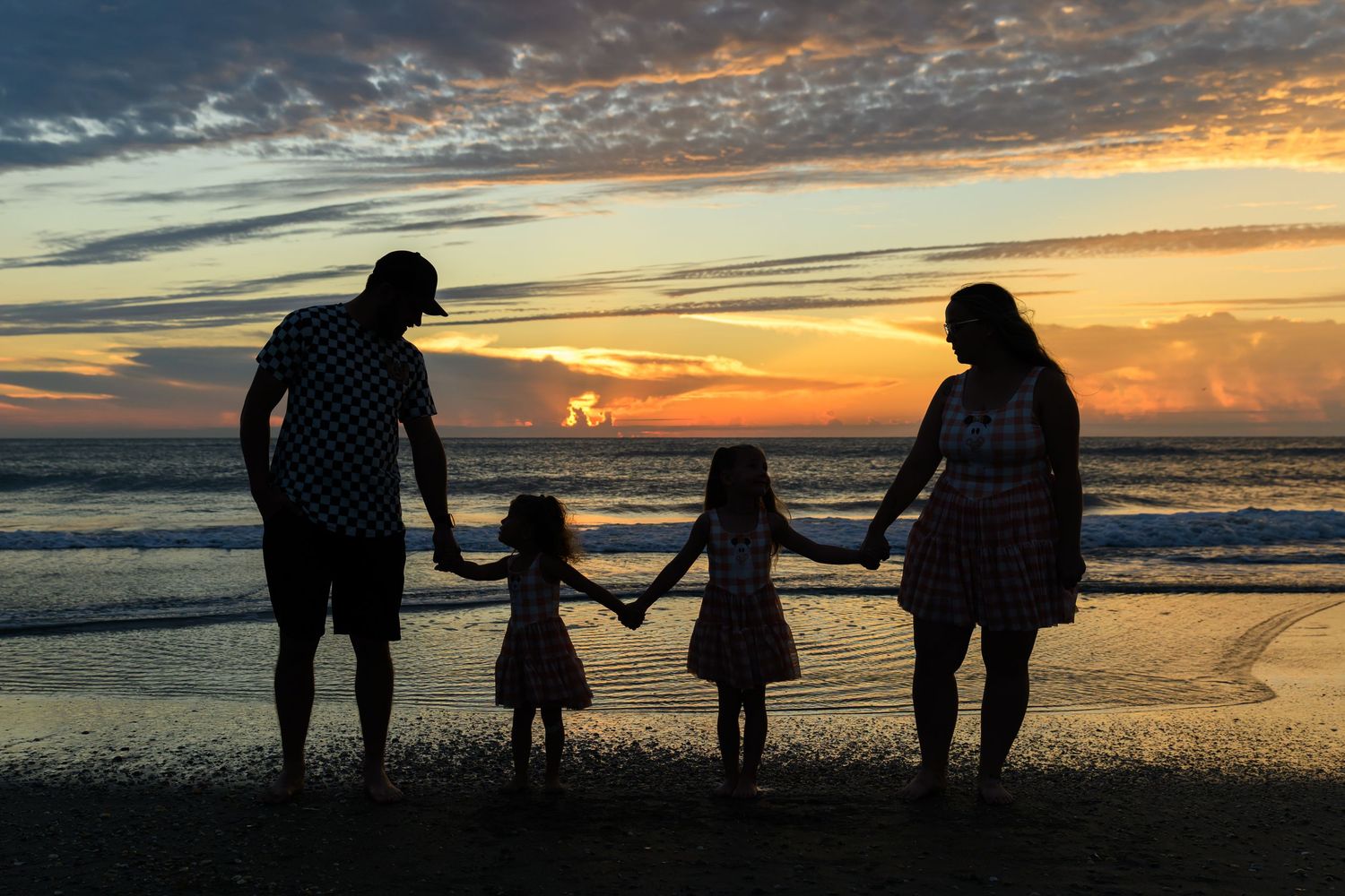 Silhouette of a family of four holding hands on a sandy beach at sunset with colorful clouds over the ocean.