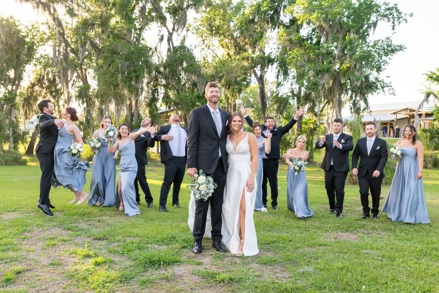 Wedding party celebrates outdoors under moss-draped trees, bride and groom in foreground, bridesmaids in blue dresses cheering.