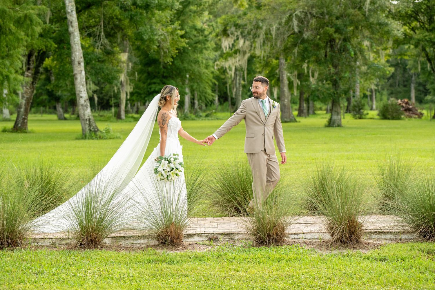 Bride and groom holding hands in lush green park, bride wearing long veil and white gown, groom in tan suit.