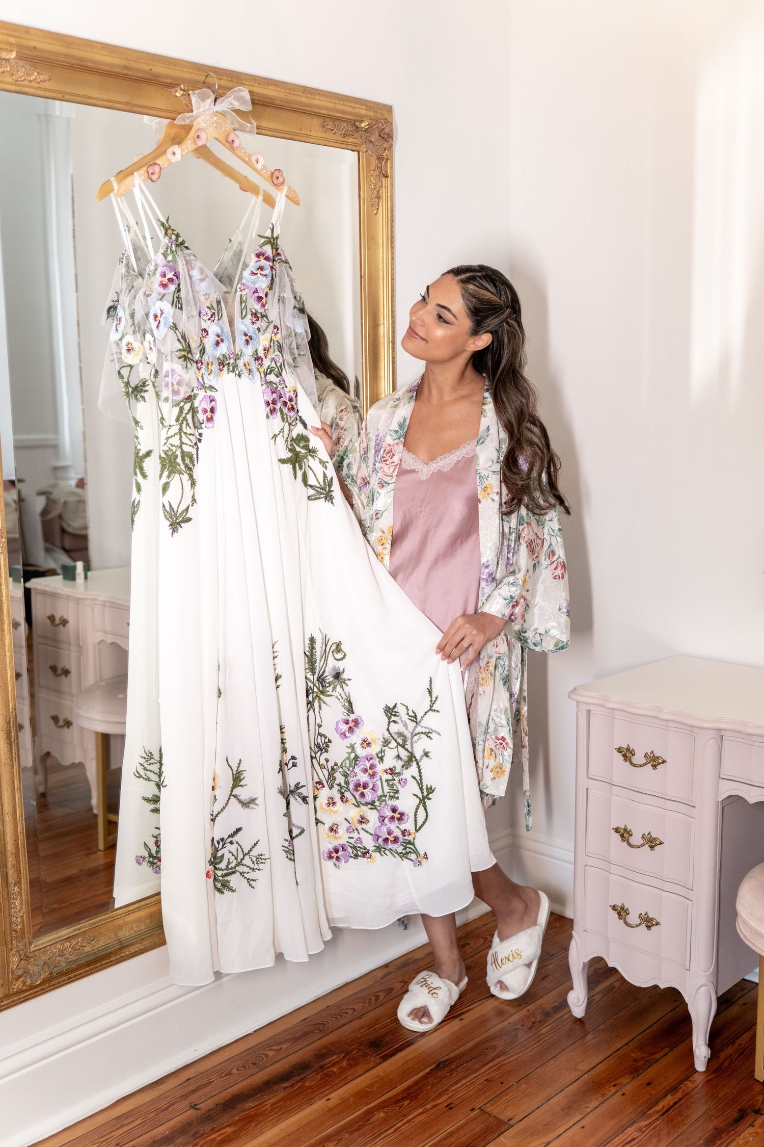 Bride in pink satin robe admiring floral wedding dress on hanger beside gold mirror and vintage dresser.