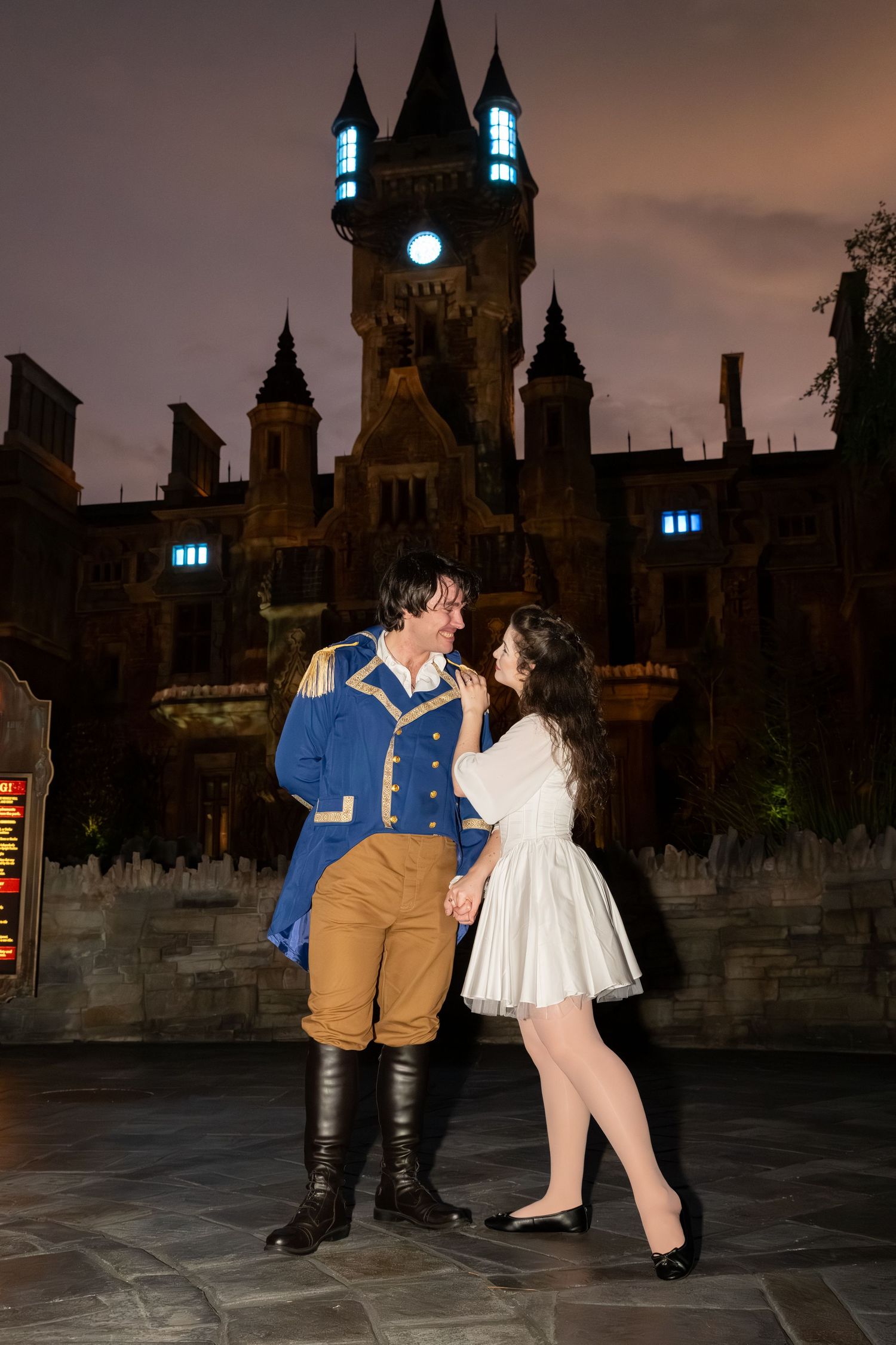 Couple in costume poses romantically before an illuminated castle at night, man in blue military jacket and woman in white dress.