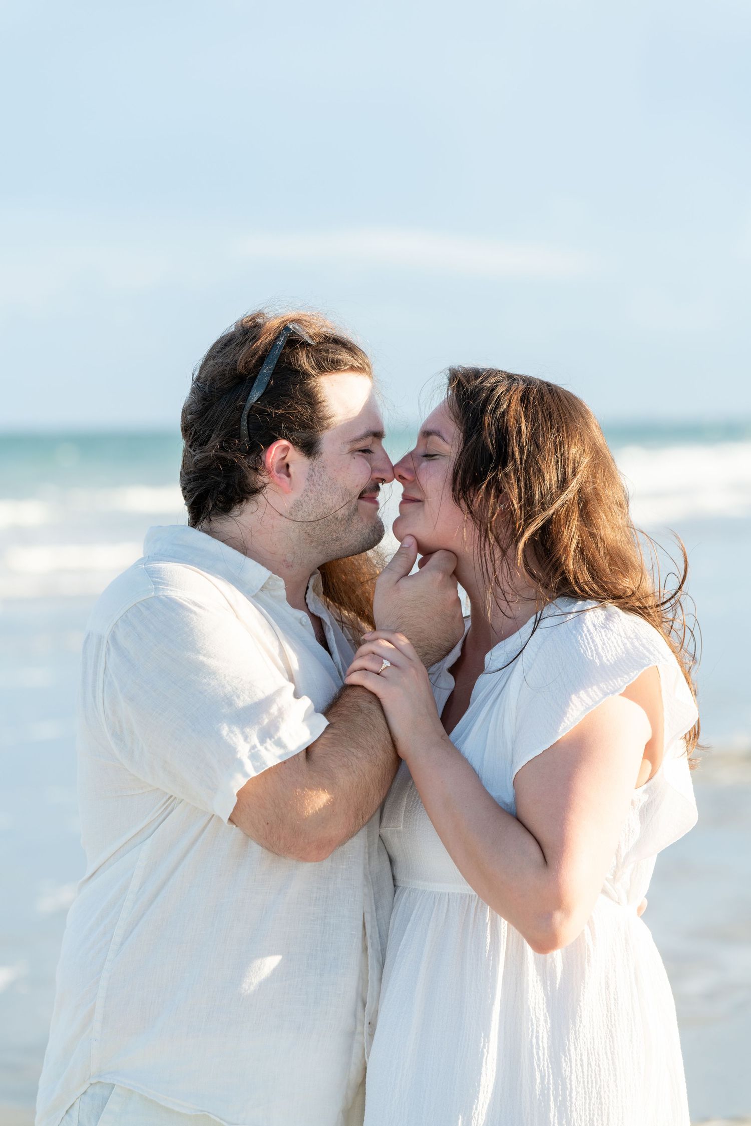 A couple in white outfits share a romantic moment on a beach, noses touching and hands clasped together.