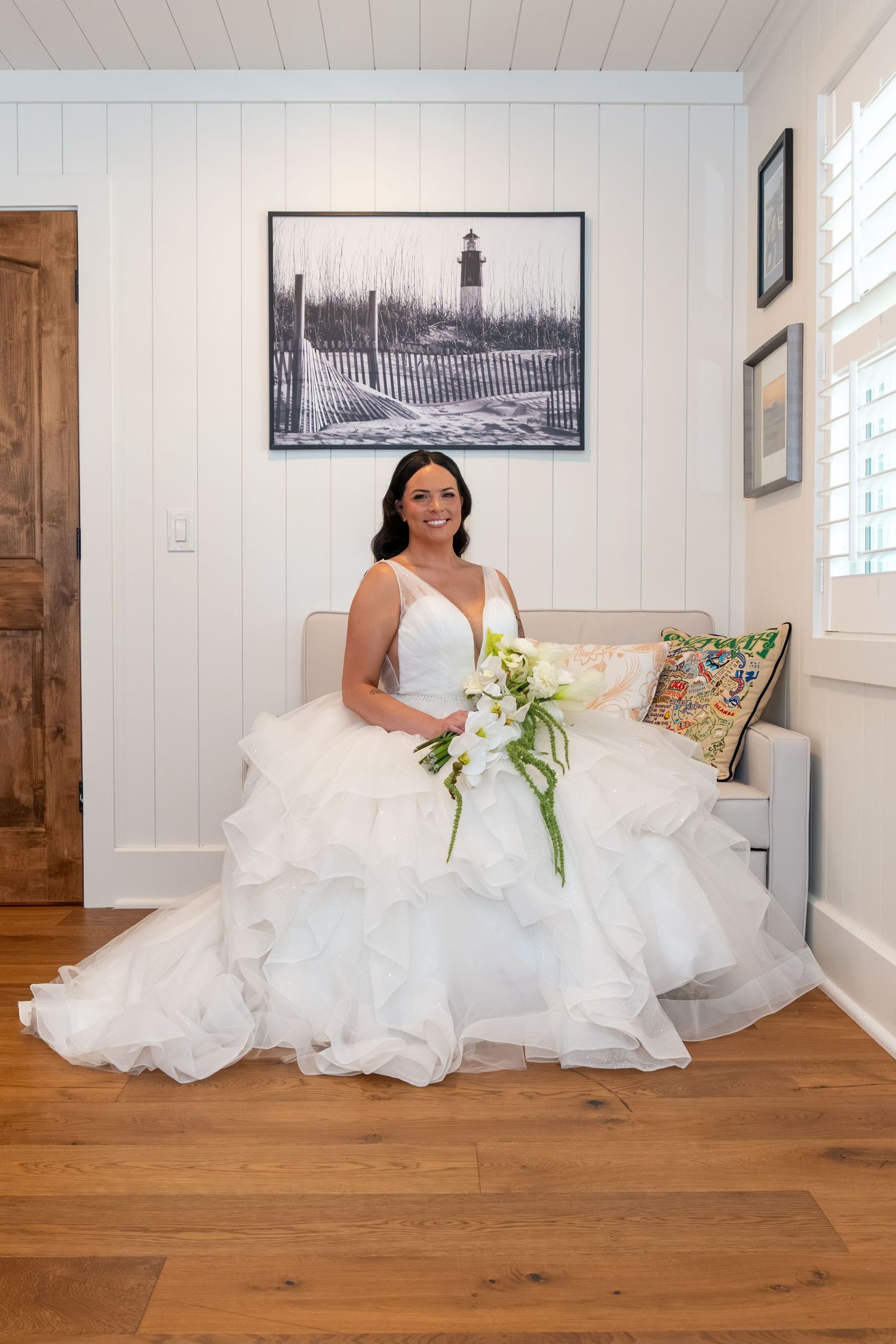 Bride in white ballgown holding white bouquet seated on couch with coastal artwork backdrop on hardwood floors.