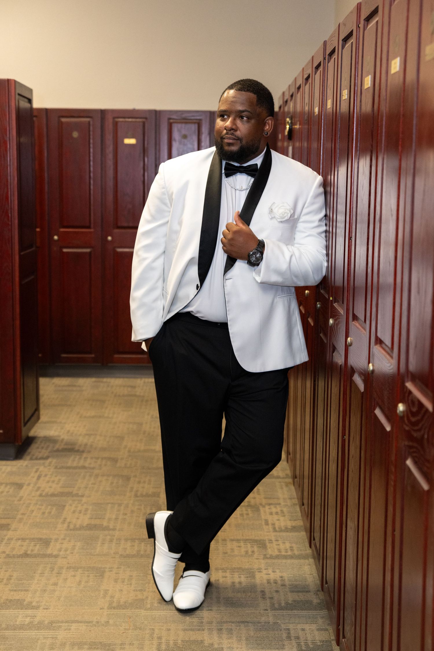 Man in white tuxedo jacket with black lapels, bow tie, and white shoes leaning against wooden lockers.