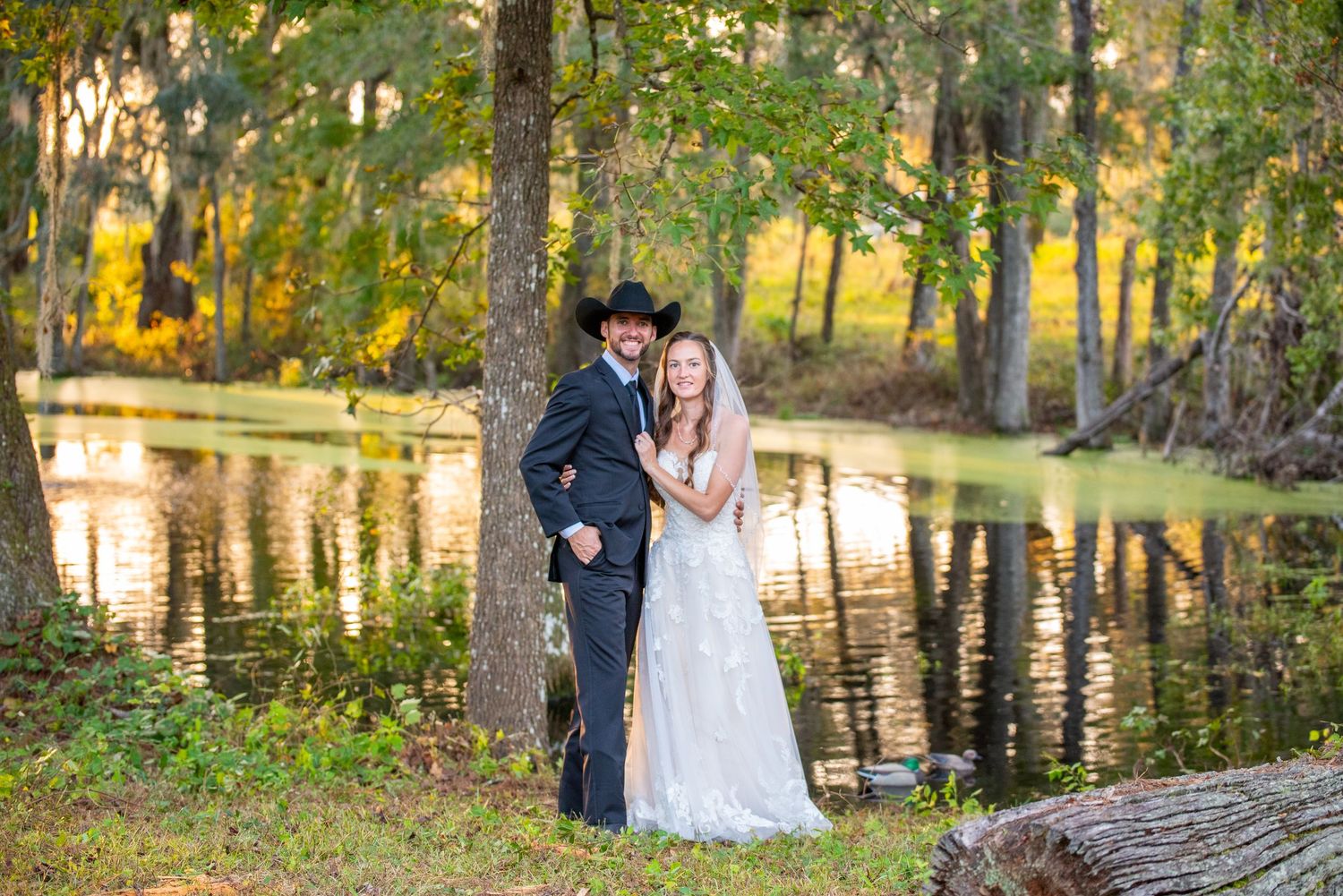 Wedding couple embraces by a serene pond surrounded by autumn foliage in the woods.