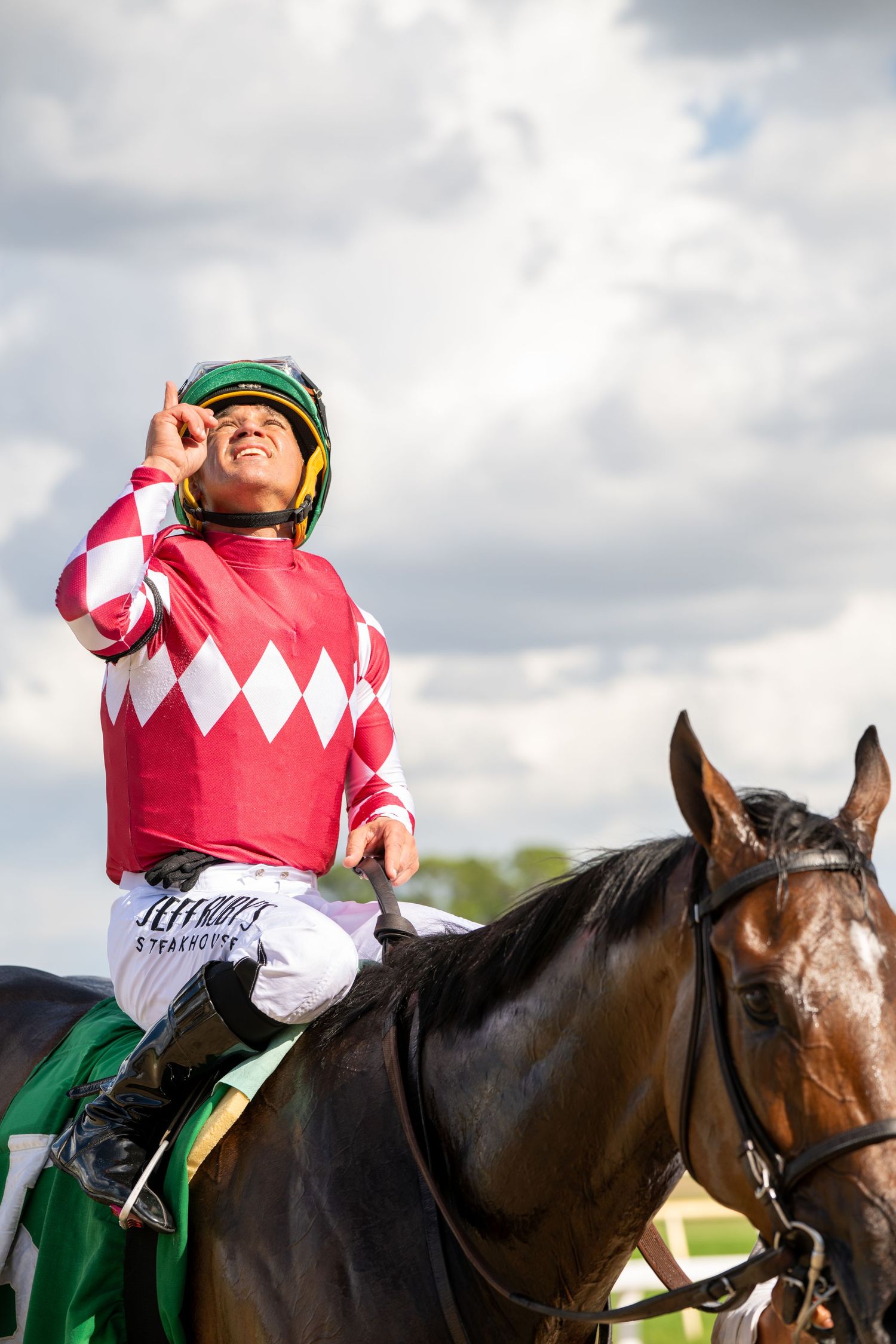 Jockey in red and white uniform celebrates victory pointing skyward while riding a dark bay horse at a horse race.